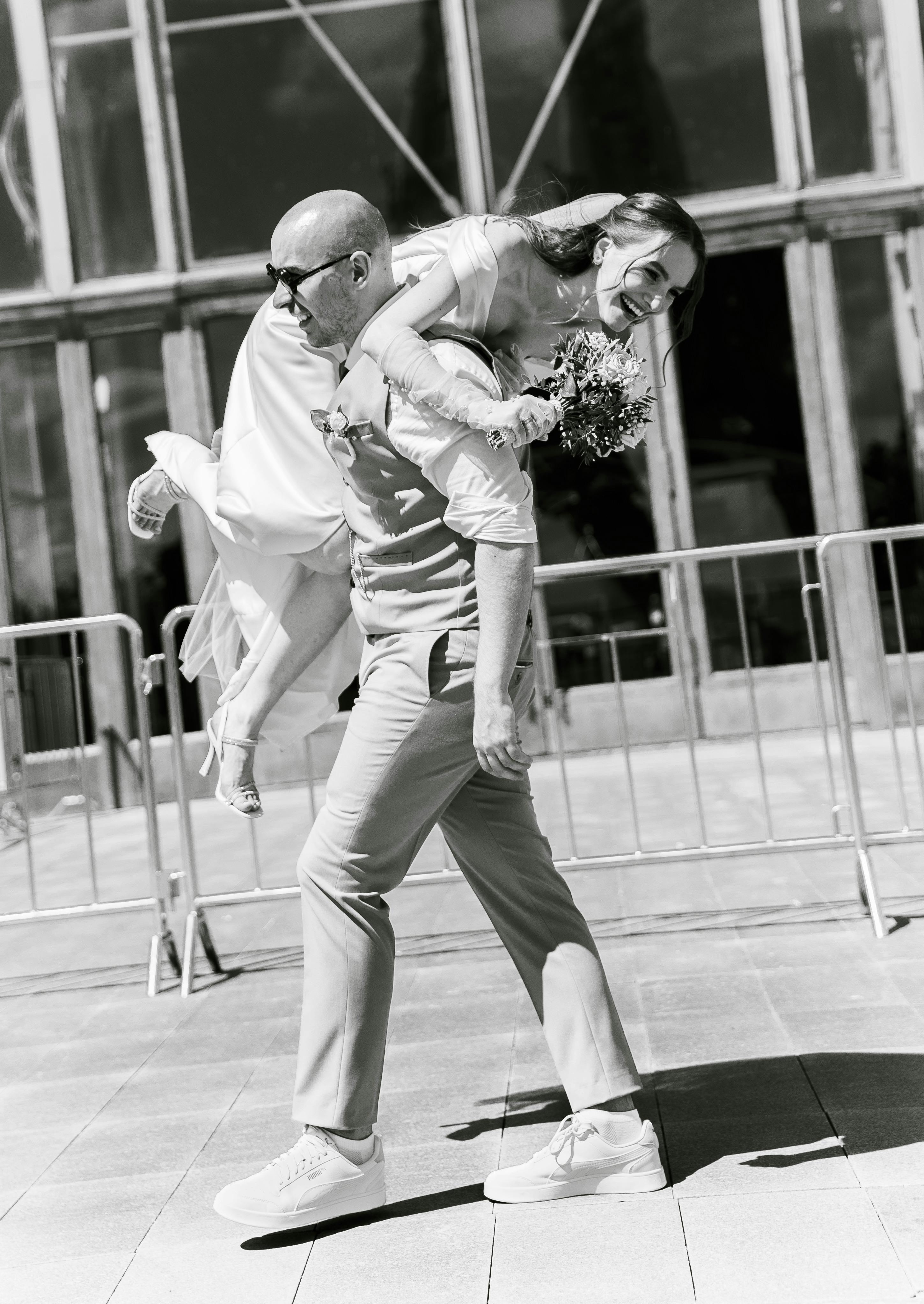 Free Bride playfully piggybacks groom in a fun outdoor wedding scene. Stock Photo