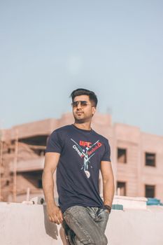 Trendy young man wearing sunglasses and casual attire leaning against a wall outdoors in summer.