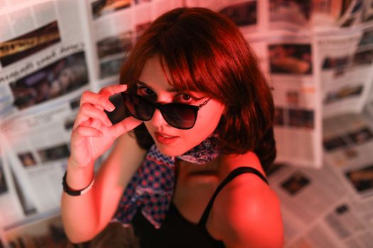 Fashionable woman wearing sunglasses with a vibrant red light, surrounded by newspapers.