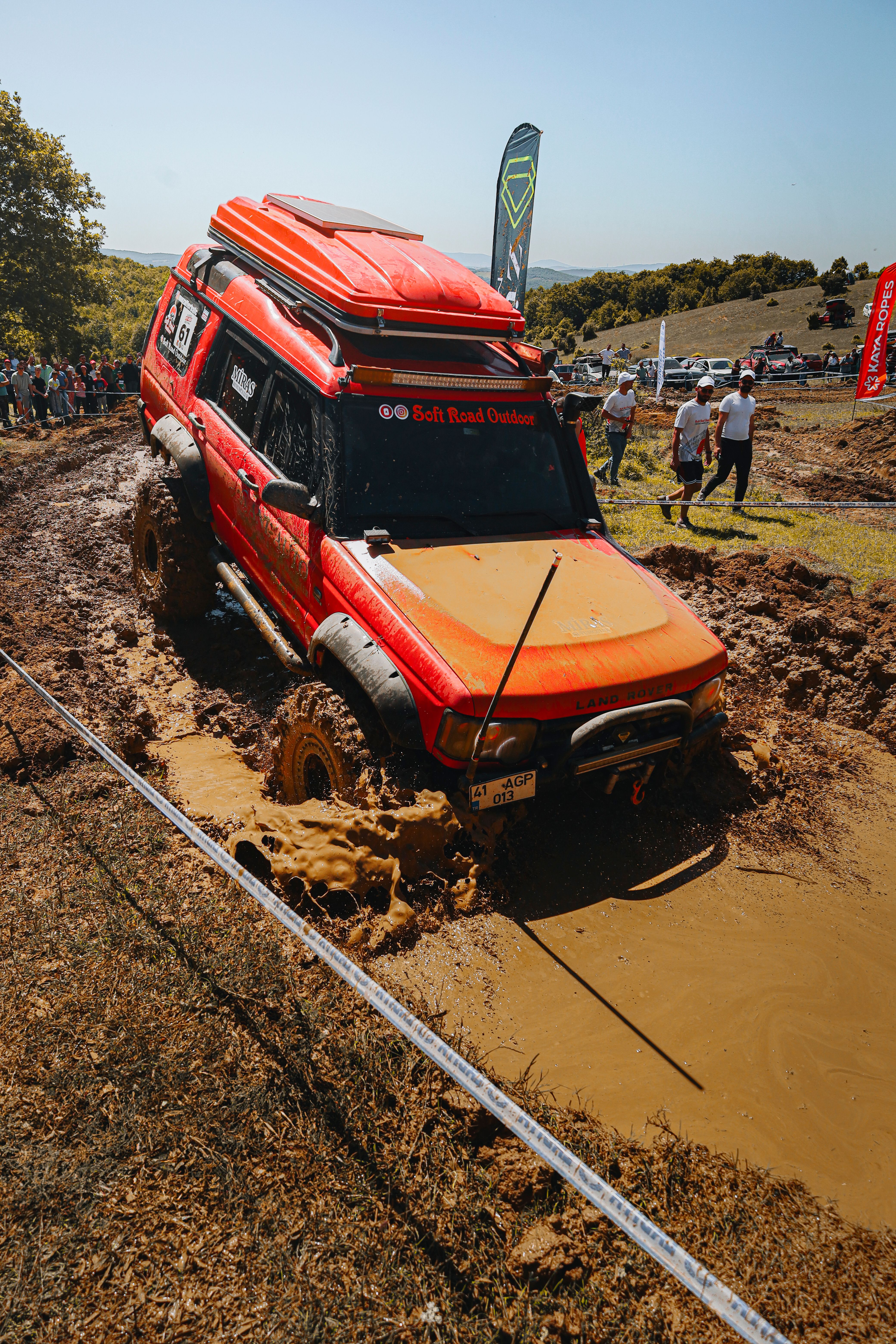 Red 4x4 SUV navigating through muddy terrain during an off-road event with spectators.