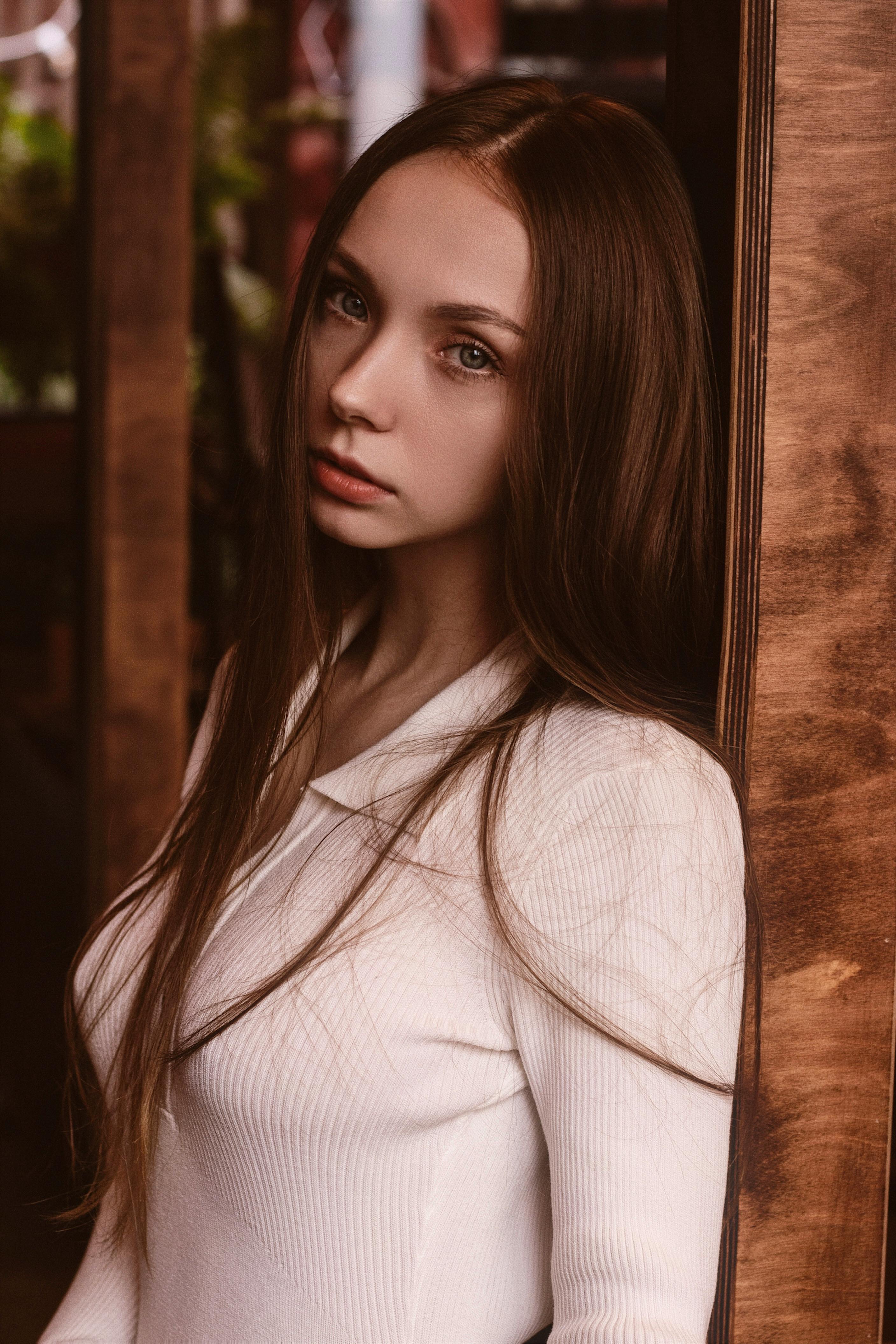 Moody portrait of a young brunette woman in a white sweater leaning against a wooden frame.