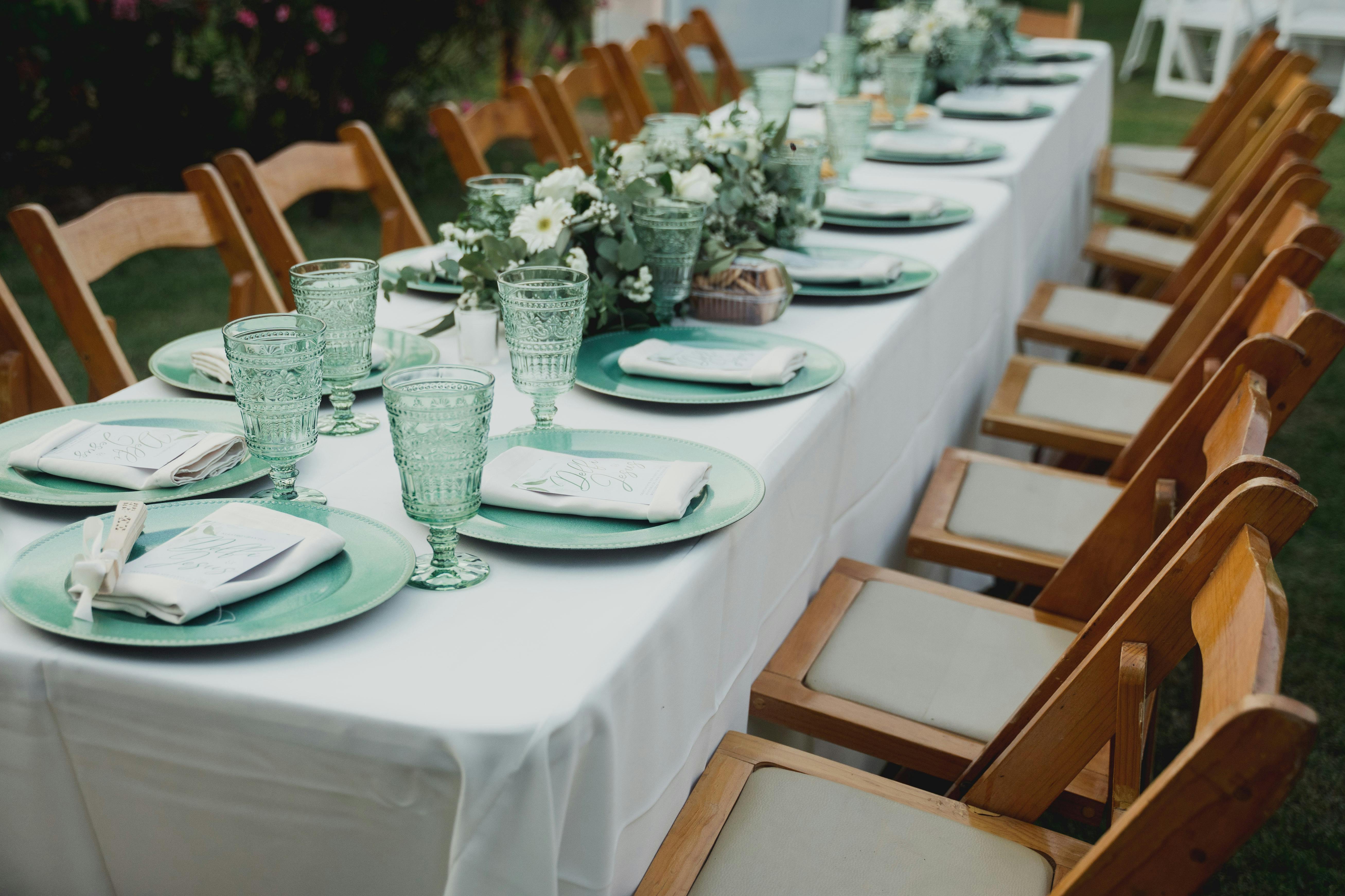 Long dining table set for an outdoor event with elegant floral centerpieces and green tableware.