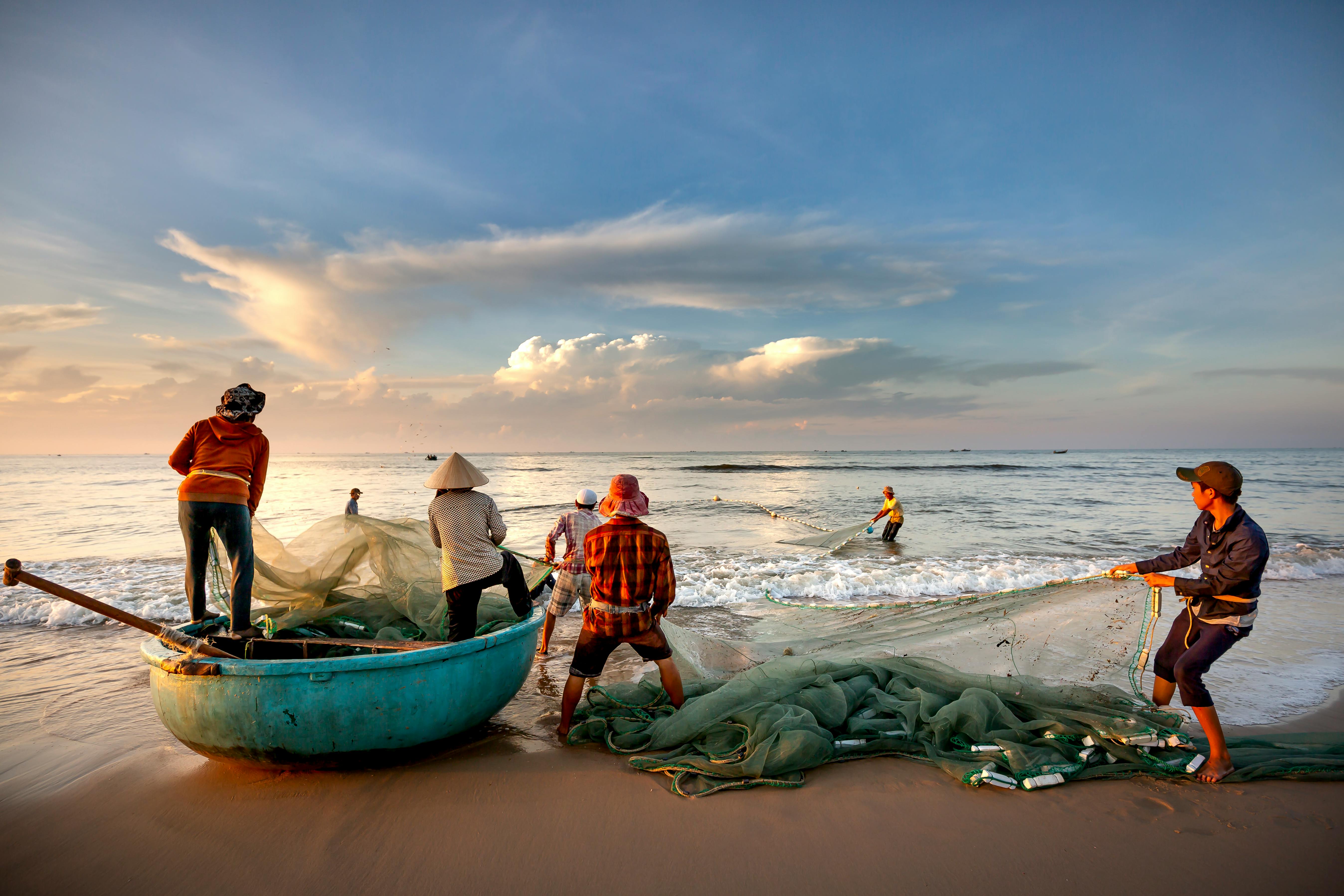 A group of diverse fishermen collaborating to haul nets on a serene beach during sunset.