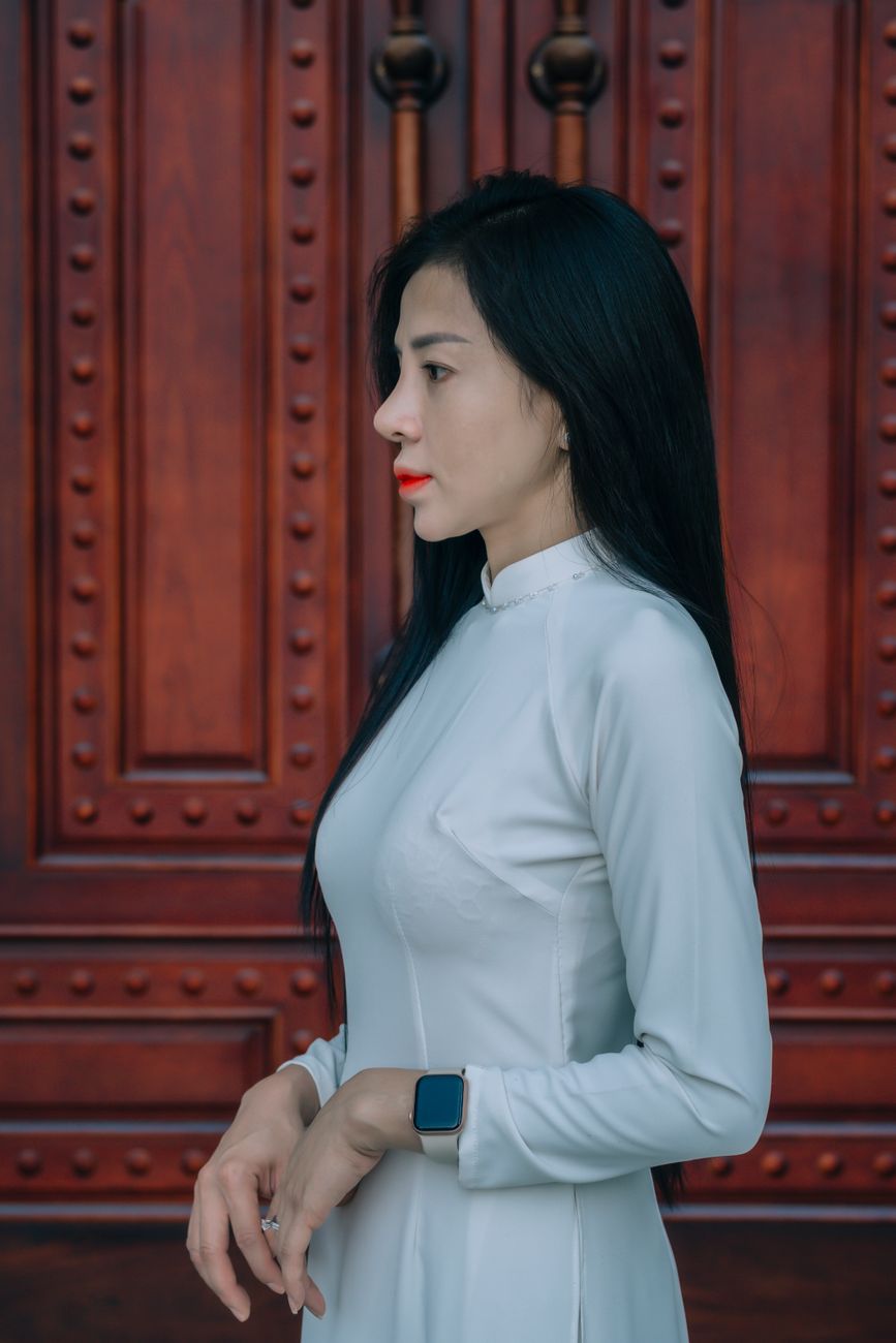 Side profile of a woman in traditional white Ao Dai dress standing by an ornate wooden door, exuding elegance.