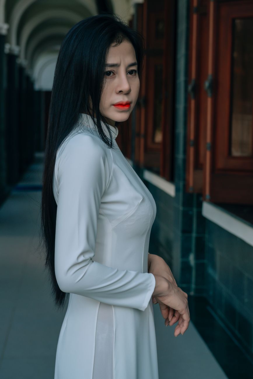 Portrait of a woman in a white Ao Dai, standing indoors with a thoughtful expression.