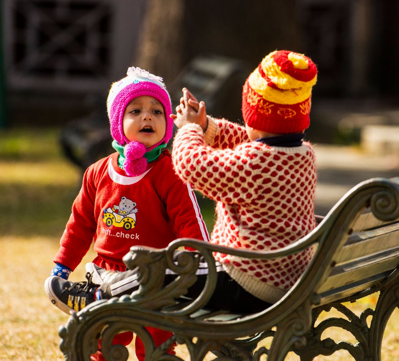 Two toddlers in colorful knit hats and sweaters sitting together on a park bench
