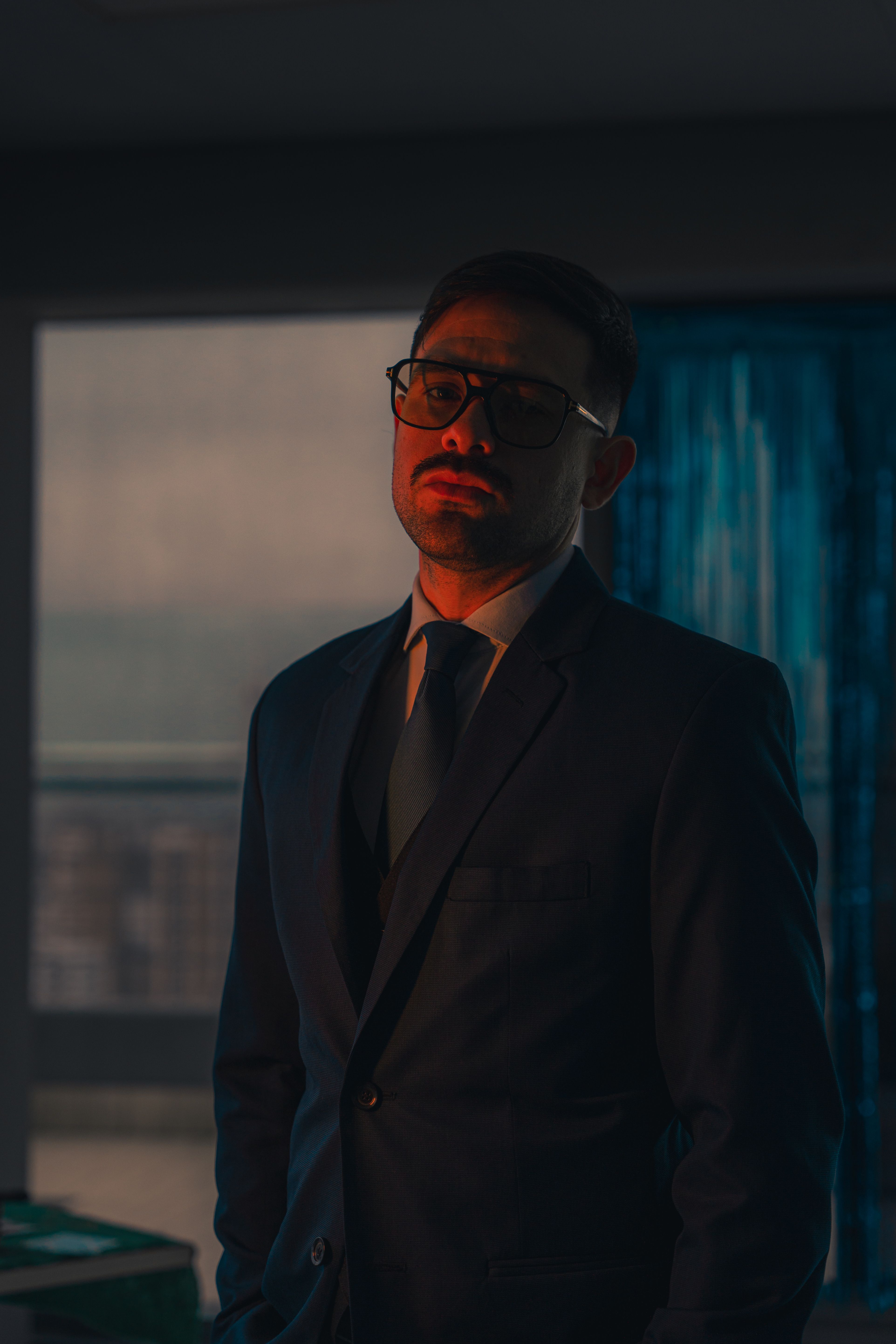 Elegant man in a suit and glasses poses with confidence in a moody, dimly lit interior setting.