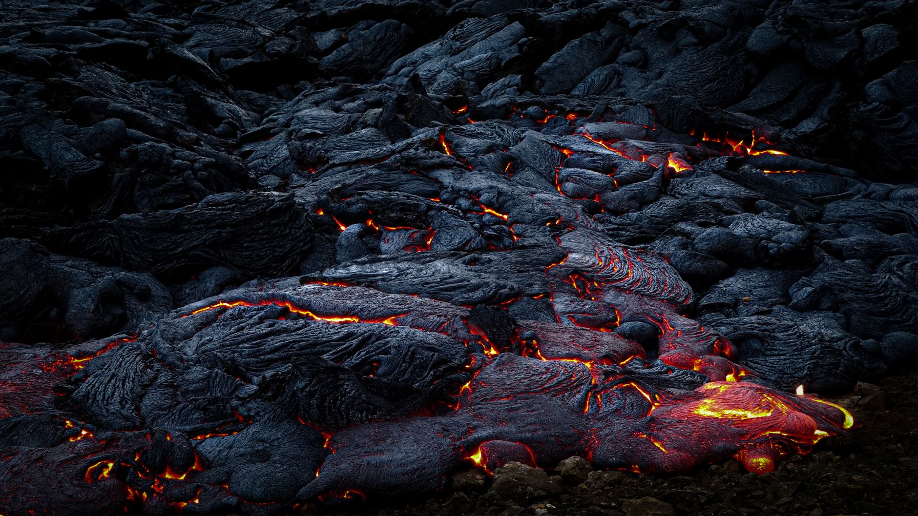 Close-up of vivid, molten lava flowing over cooled, dark volcanic rocks at night.