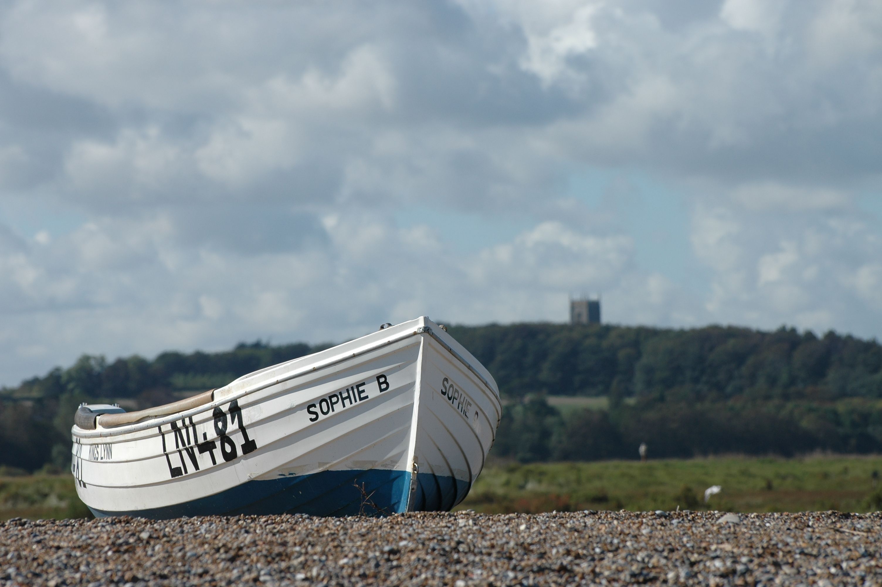 A solitary fishing boat named Sophie B rests on a pebble beach beneath a cloudy sky, conveying a sense of calm and solitude.
