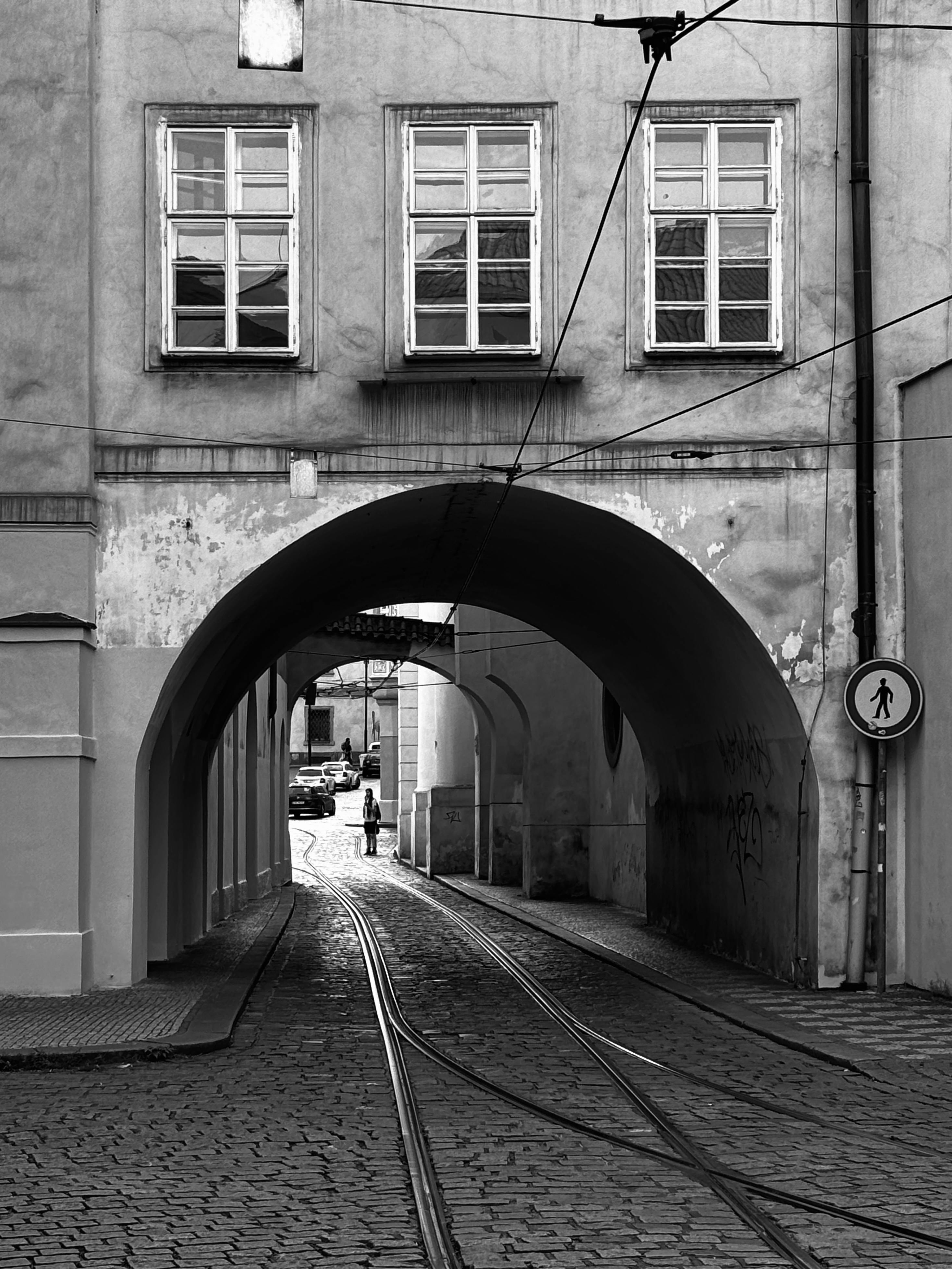 Black and white view of a historic Prague street with tram tracks under an archway.