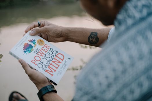 A man reads 'The Power of Your Subconscious Mind' by a riverside, showcasing relaxation and reflection.