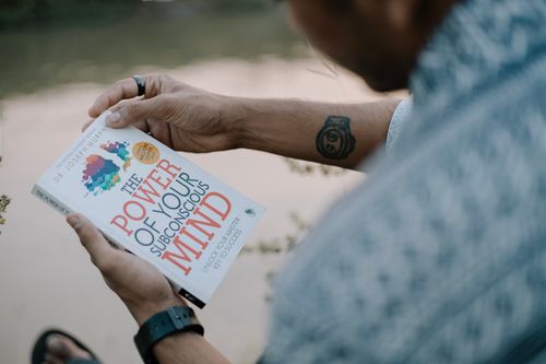 Free A man reads 'The Power of Your Subconscious Mind' by a riverside, showcasing relaxation and reflection. Stock Photo