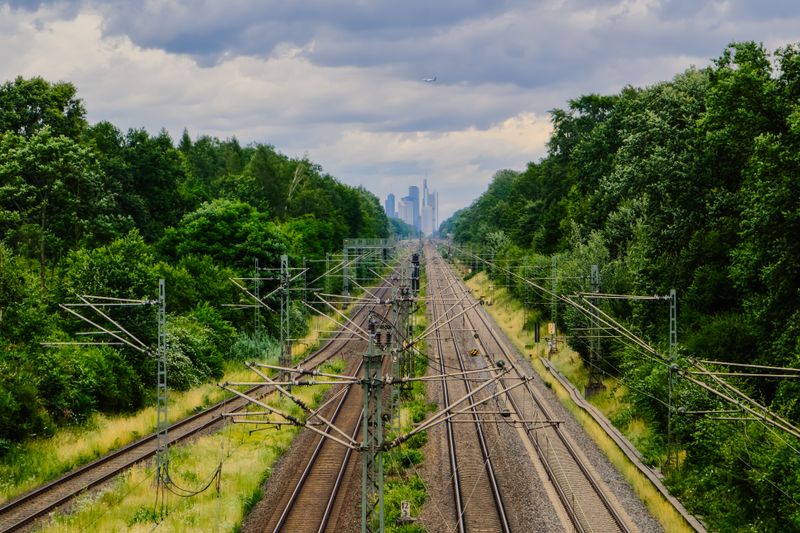 Railway tracks stretch through lush greenery towards a distant urban skyline under a cloudy sky.