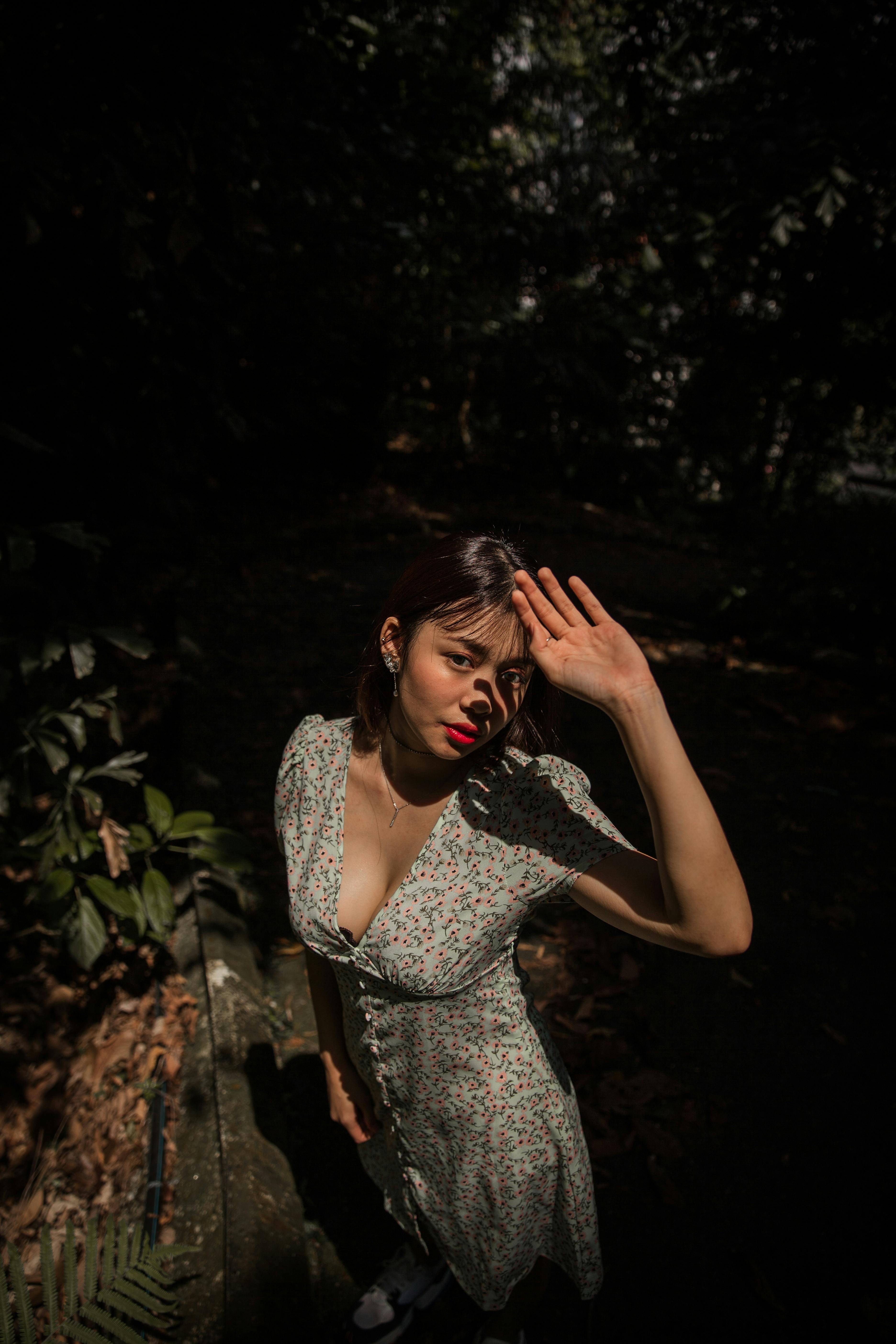 A young woman in a floral dress poses outdoors, shielding her eyes from the sunlight.