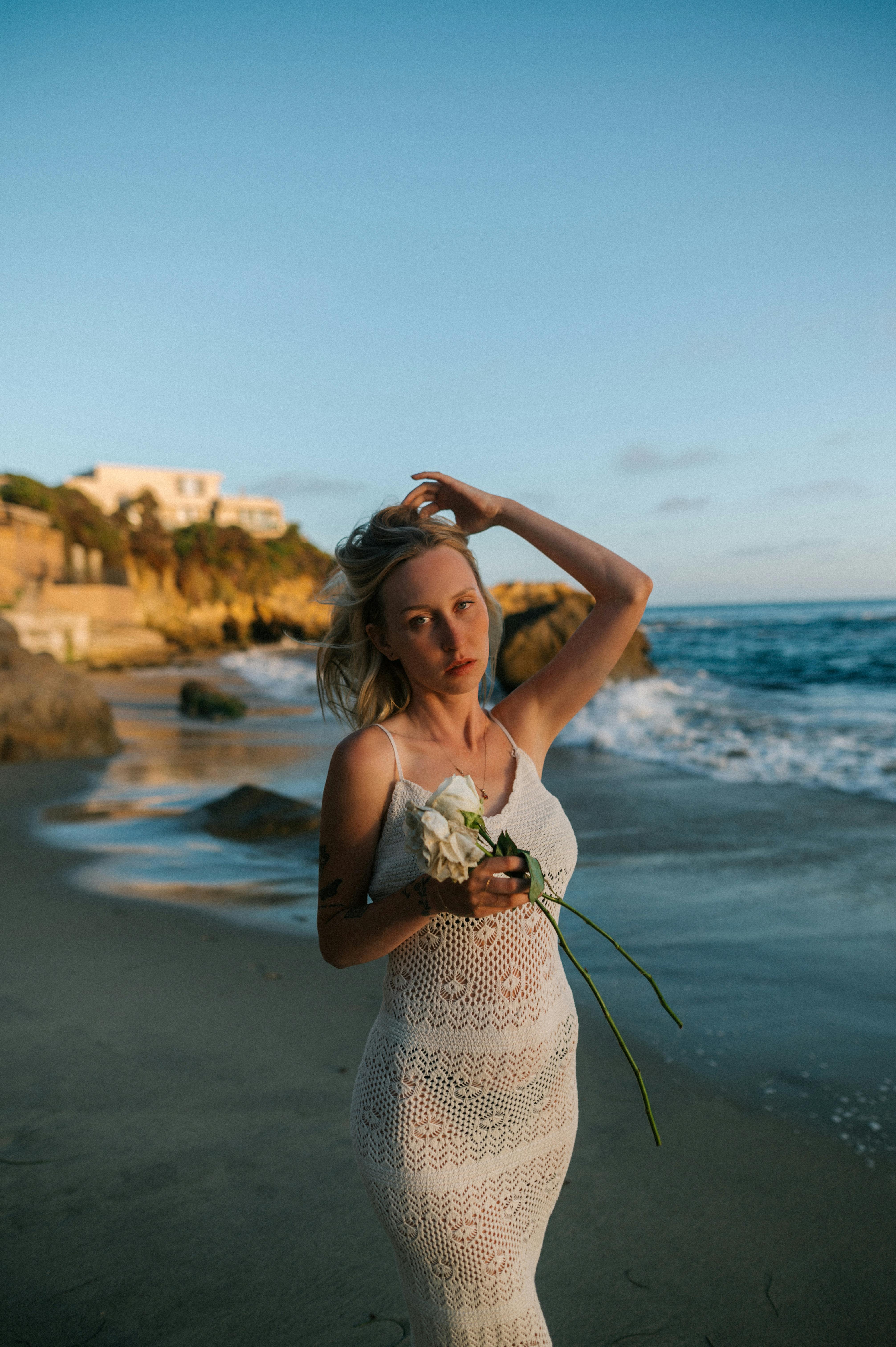 A woman holds flowers as she stands on a picturesque beach at sunset, wearing a white dress.
