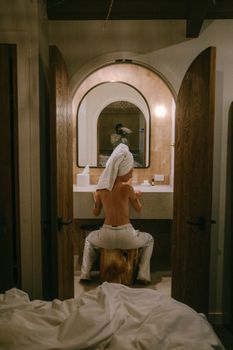 Woman with towel relaxes in front of a mirror in a cozy bedroom setting.