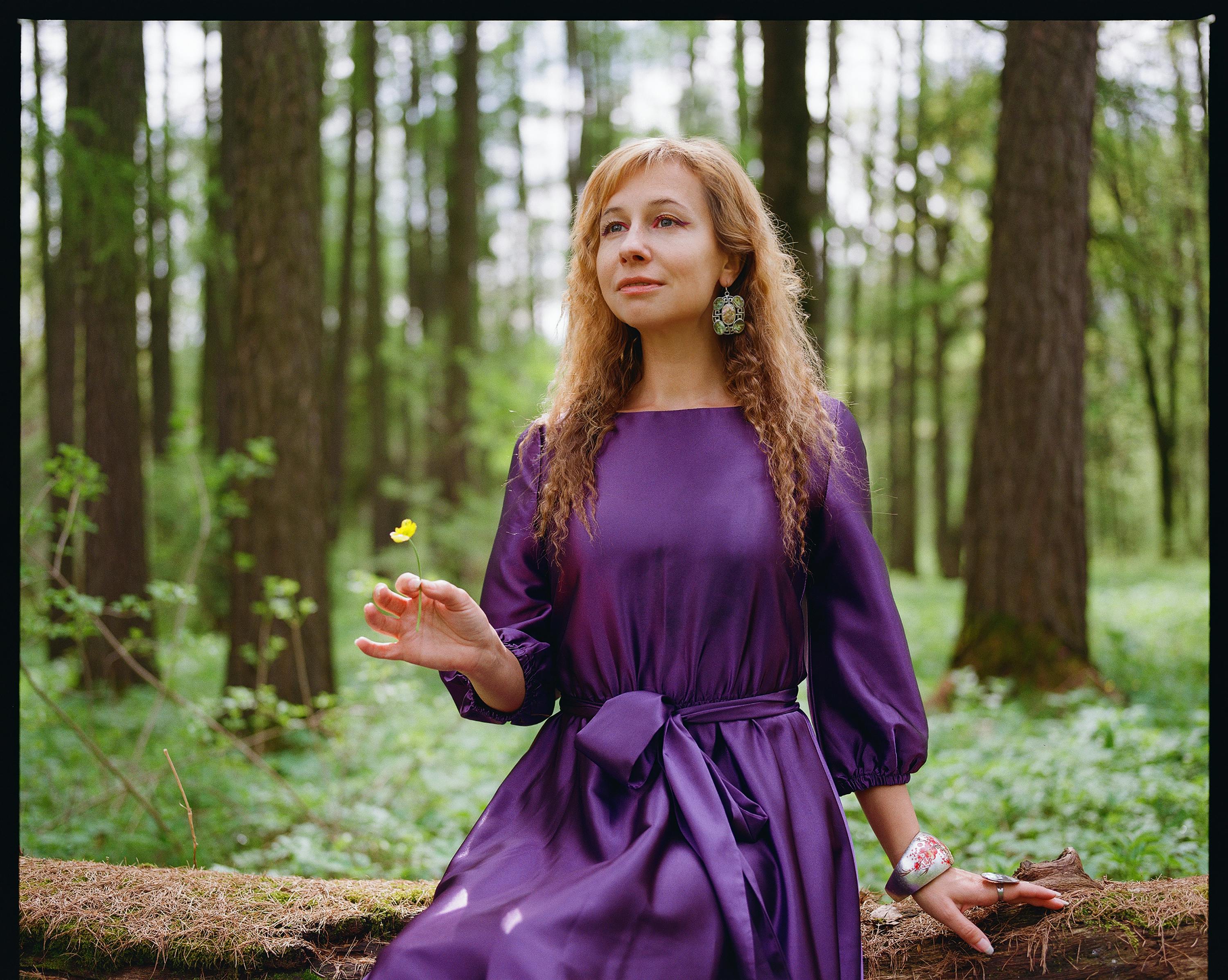 A serene portrait of a woman in a purple dress sitting in a lush green forest during daytime.