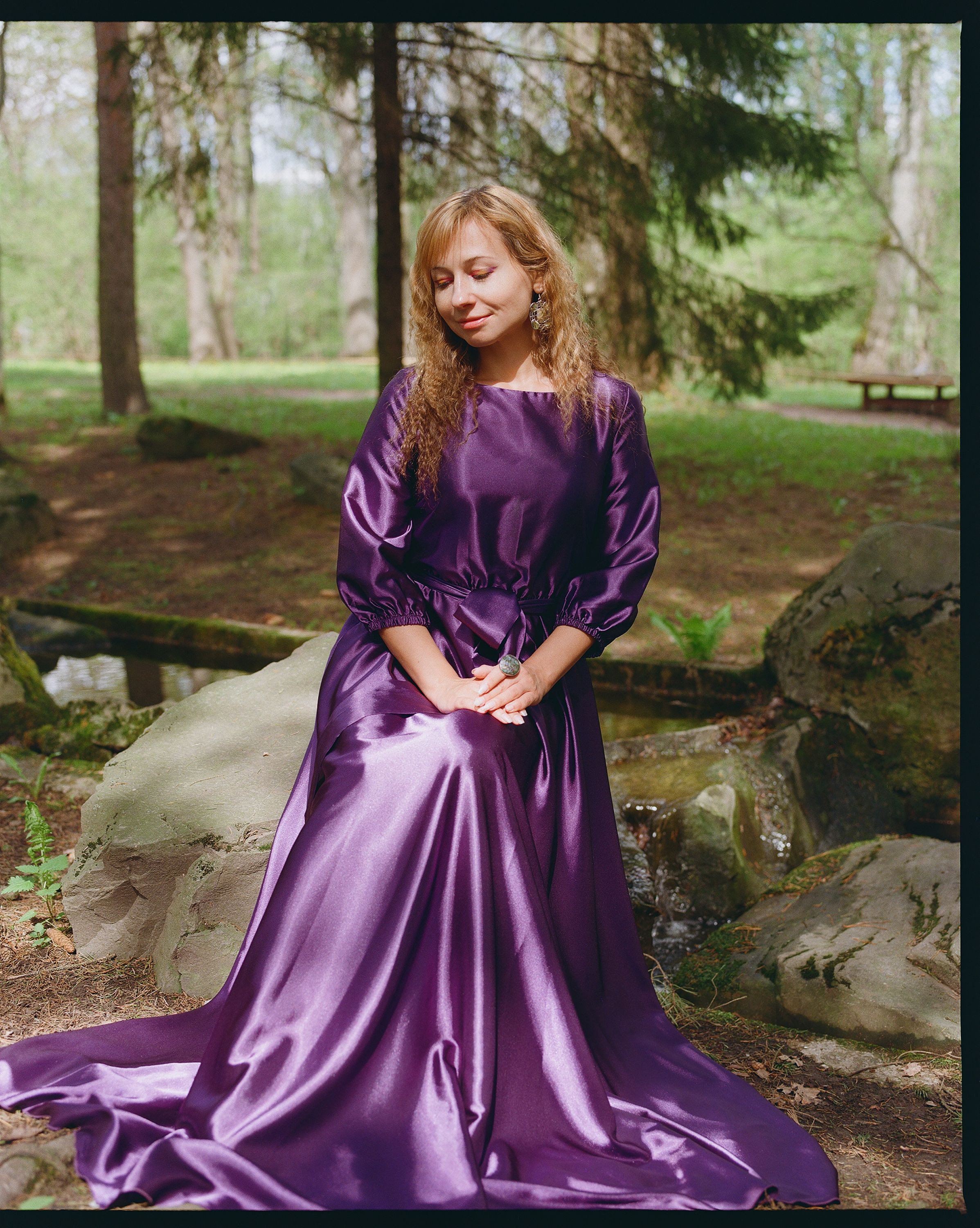 A woman in a beautiful purple gown sitting in a serene forest environment.