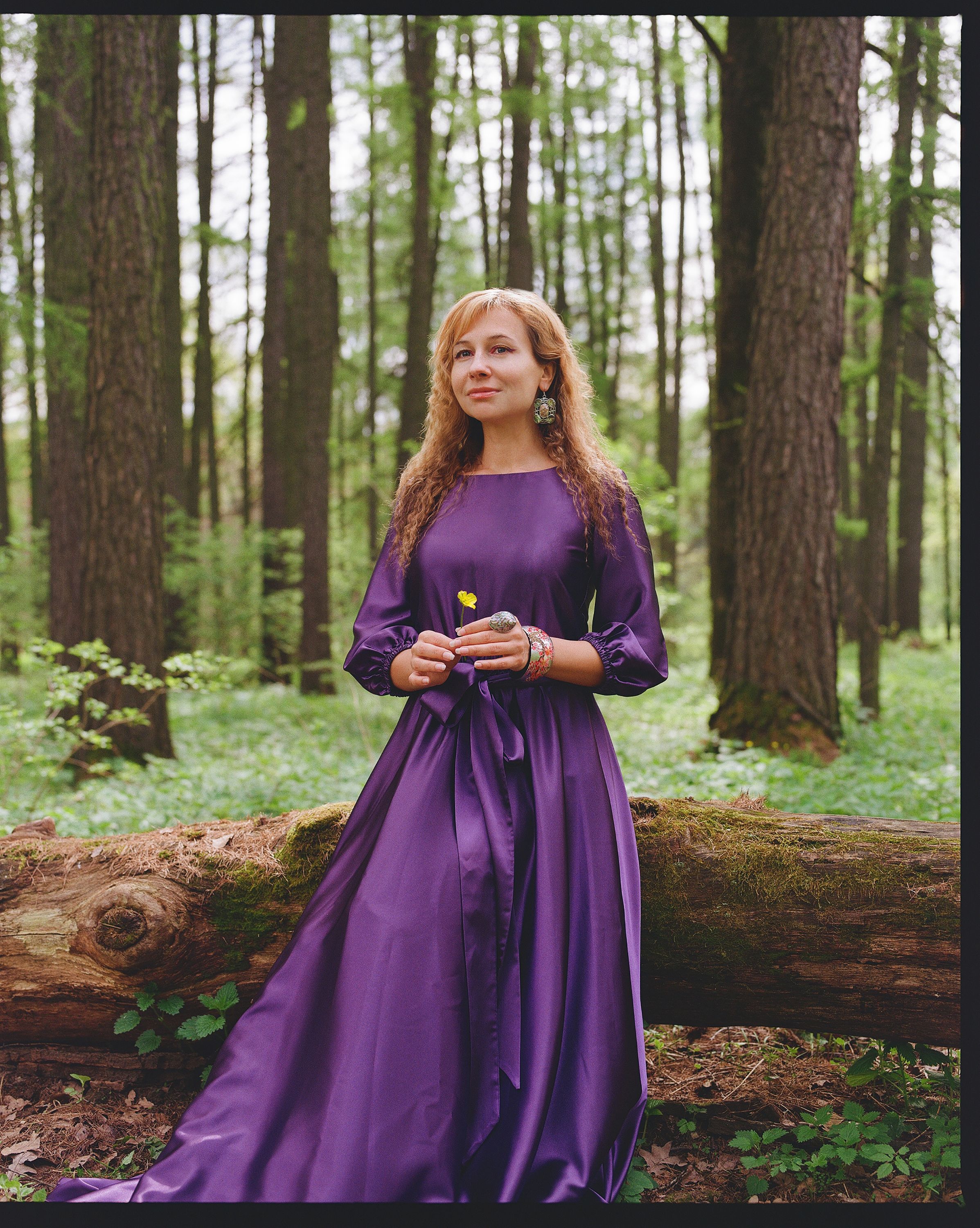 A serene portrait of a woman in a purple dress sitting on a log in a lush green forest.