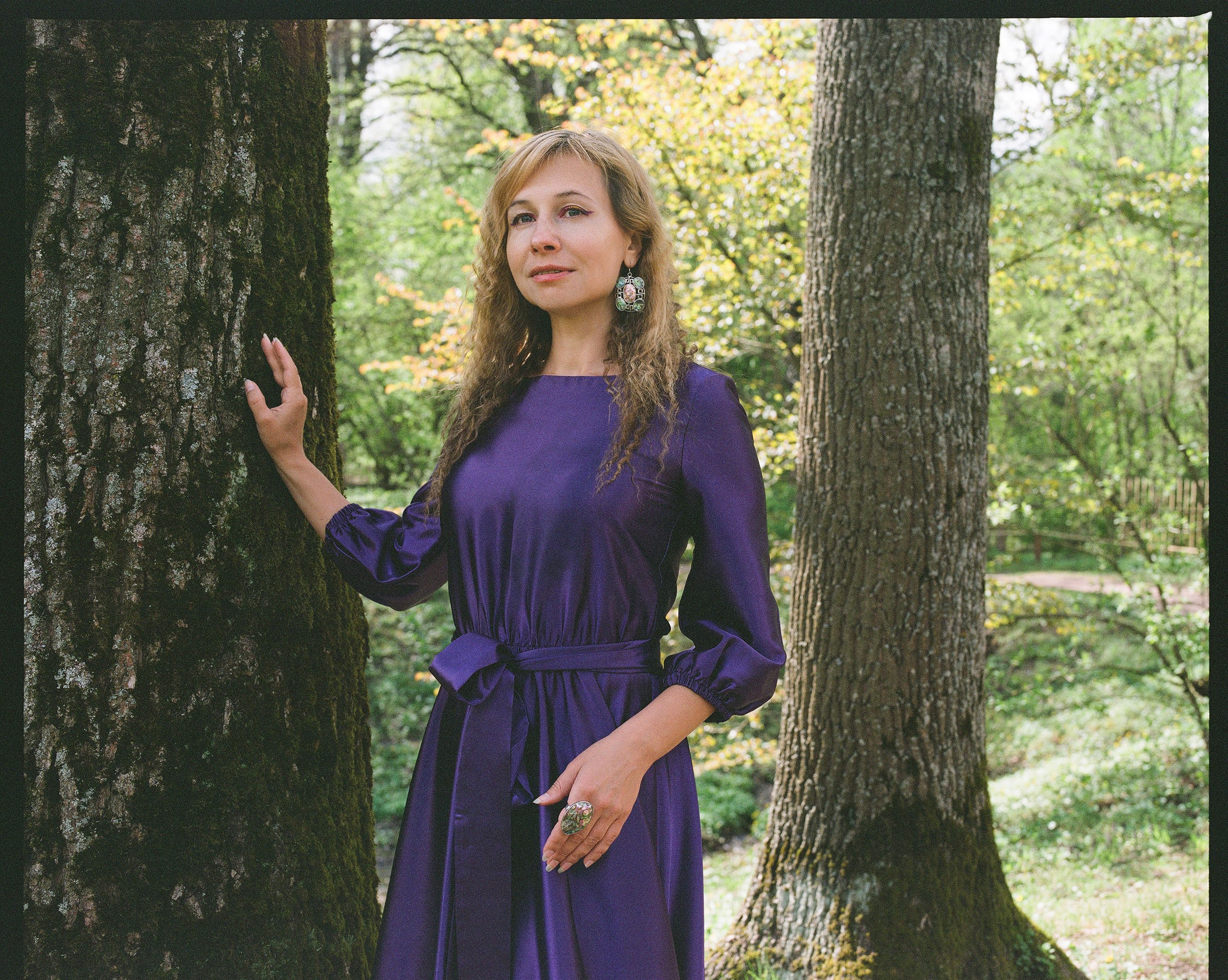 Portrait of an elegant woman in a purple dress posing in a vibrant green park, showcasing seasonal fashion.
