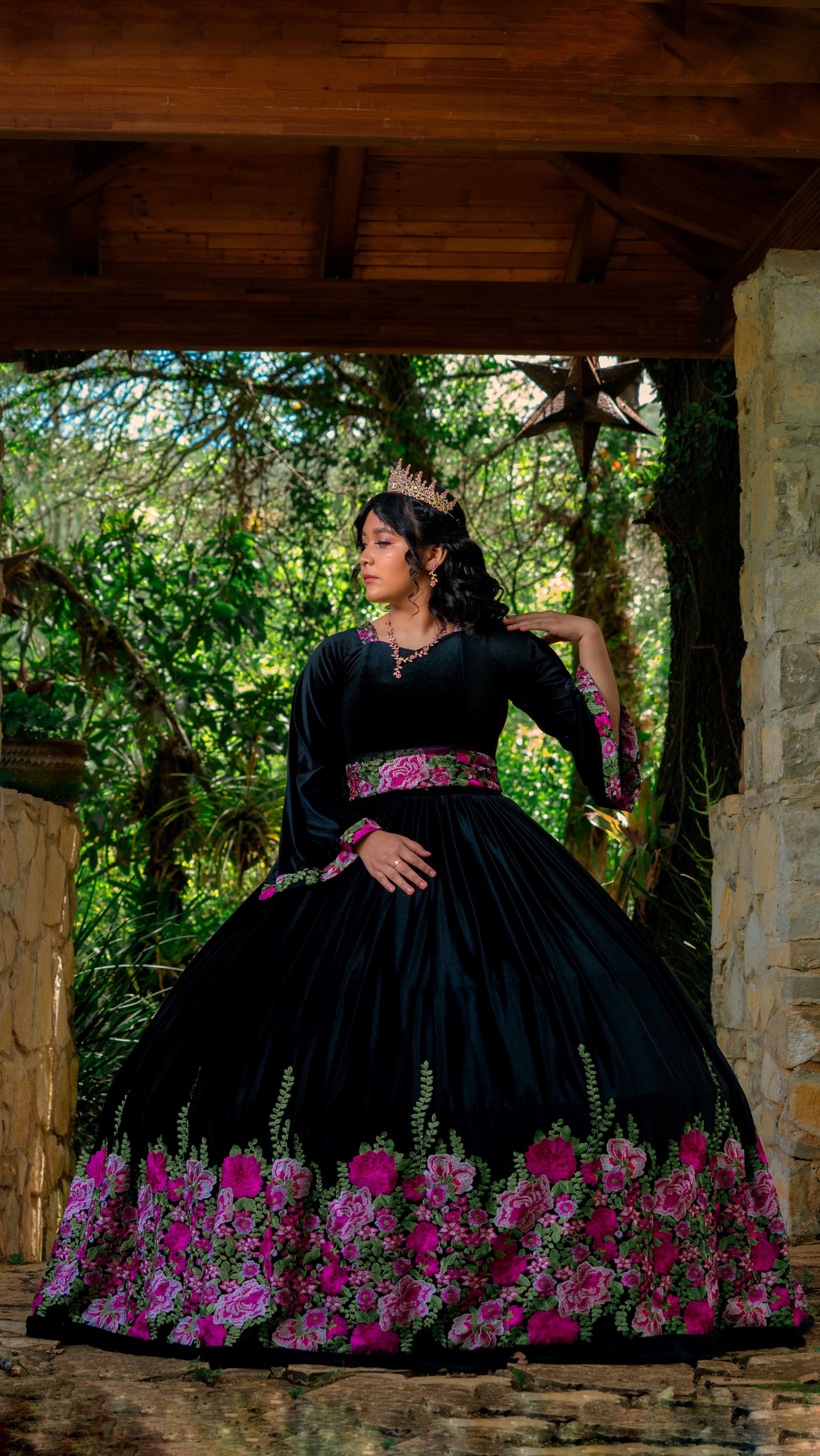A woman in a black gown with floral embroidery poses elegantly outdoors surrounded by nature.