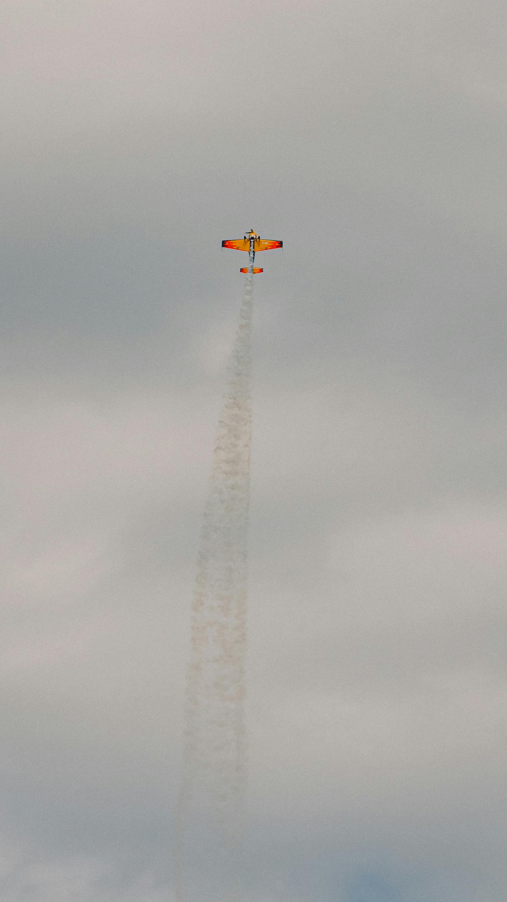 A vivid orange stunt plane ascends through clouds, leaving a trail of smoke, showcasing aerial acrobatics.