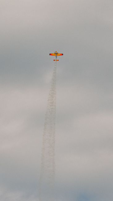A vivid orange stunt plane ascends through clouds, leaving a trail of smoke, showcasing aerial acrobatics.