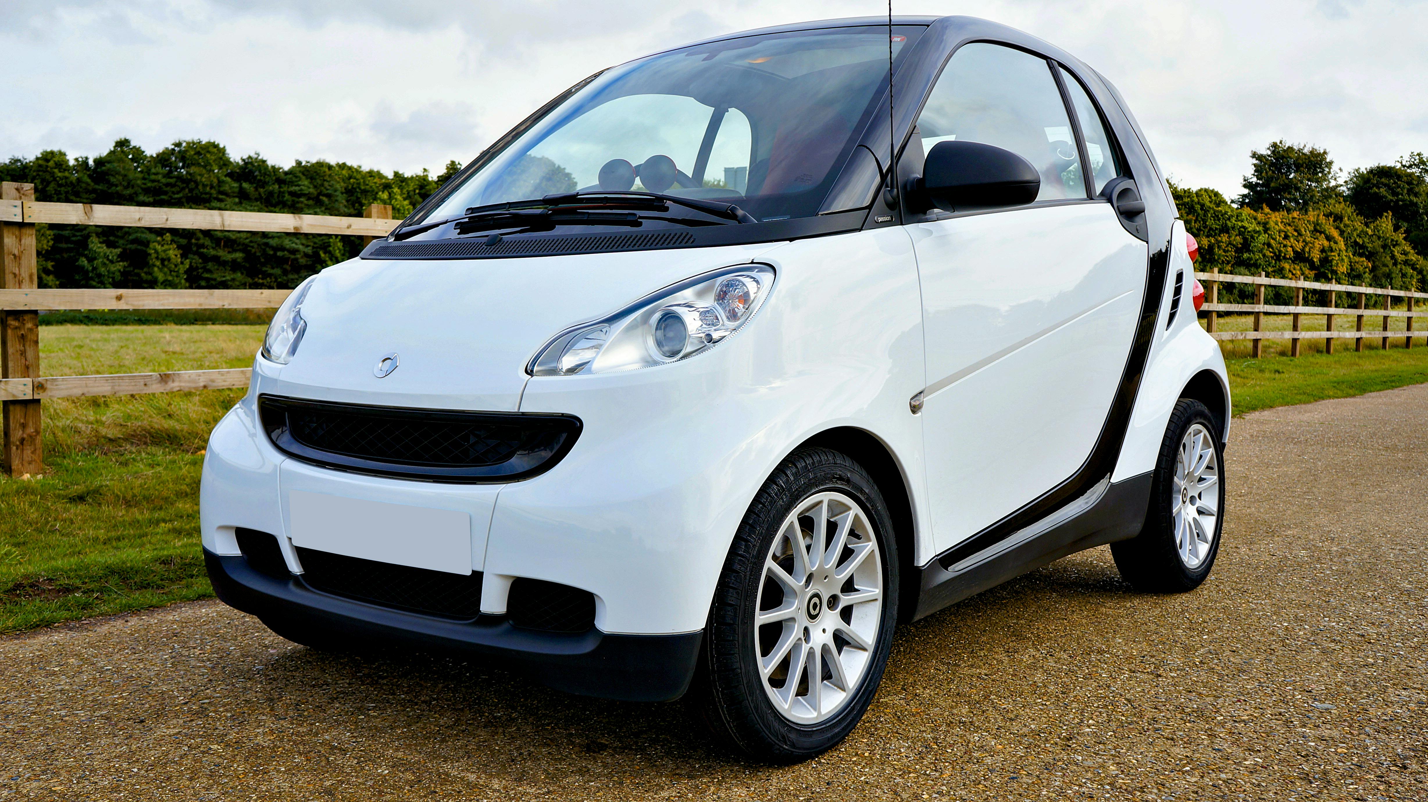 A white Smart Fortwo car parked outdoors on a sunny day with greenery and wooden fence.
