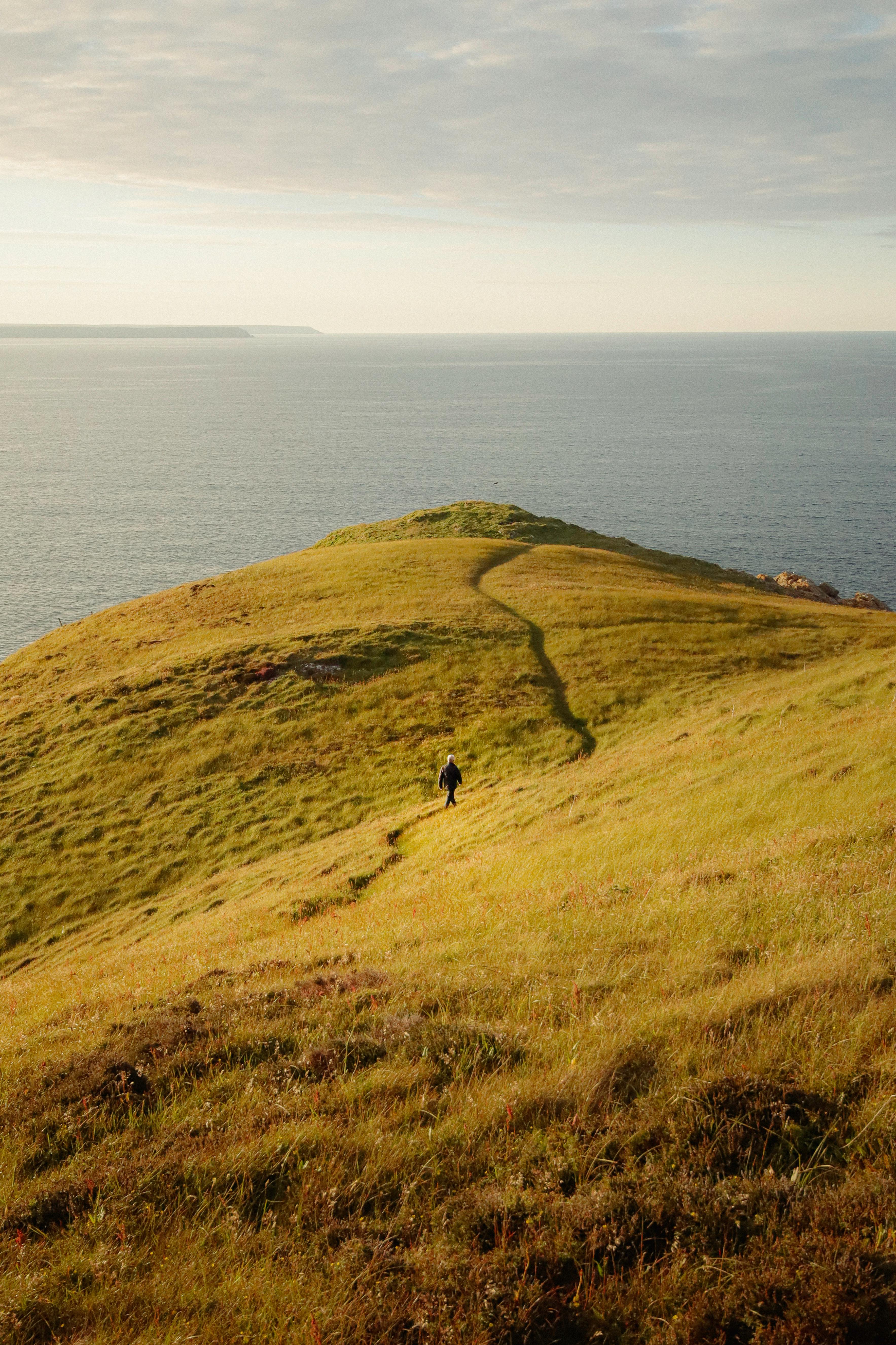 A lone hiker walks along a grassy path on a coastal hill overlooking the ocean at sunset.