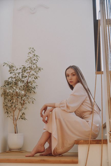 A stylish woman sits barefoot on stairs next to a potted plant, capturing a serene fashion moment.