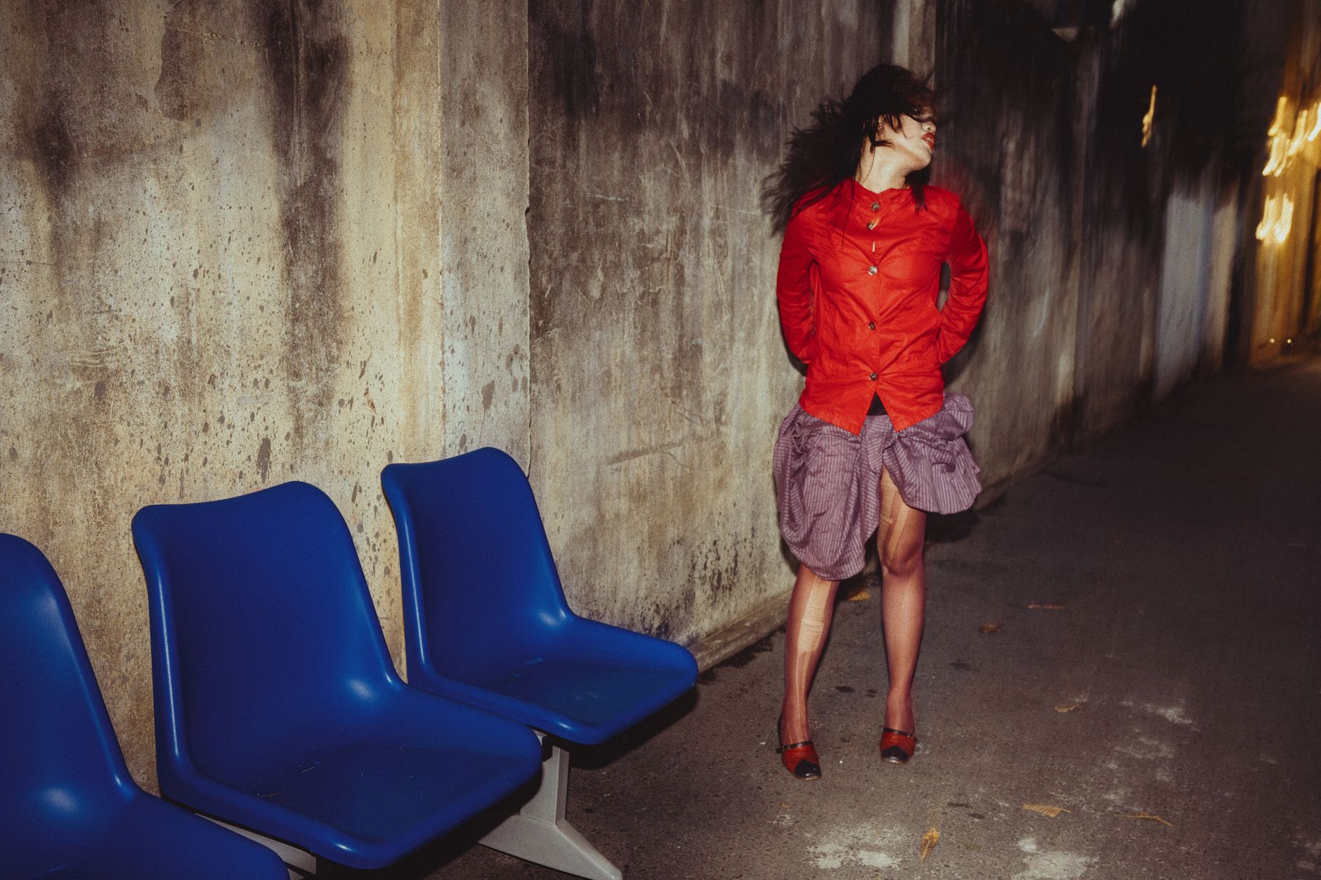 Woman in vibrant urban setting with blue chairs, red shirt, and graffiti wall.