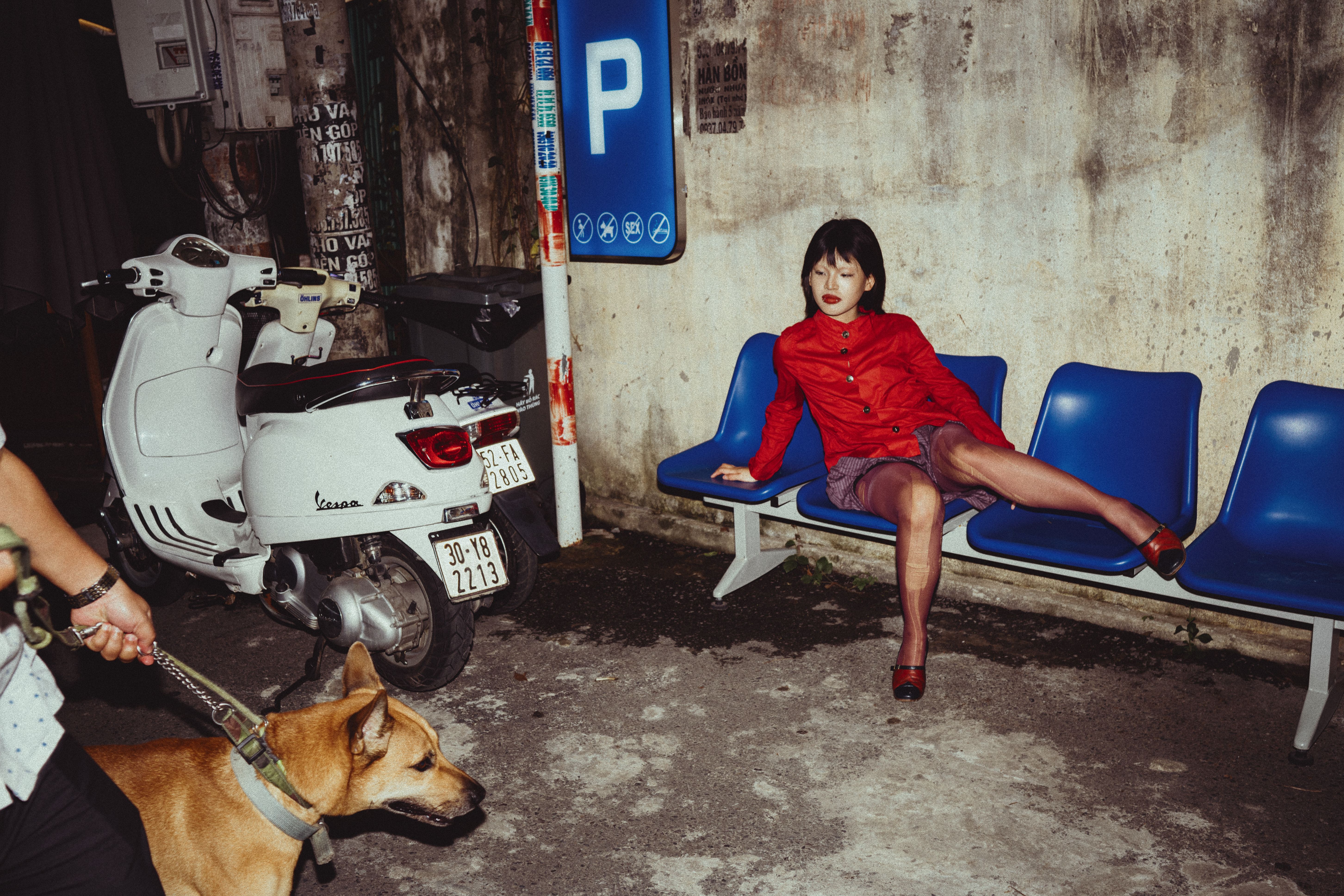 A woman in a red coat sits on a bench with a scooter nearby at night.