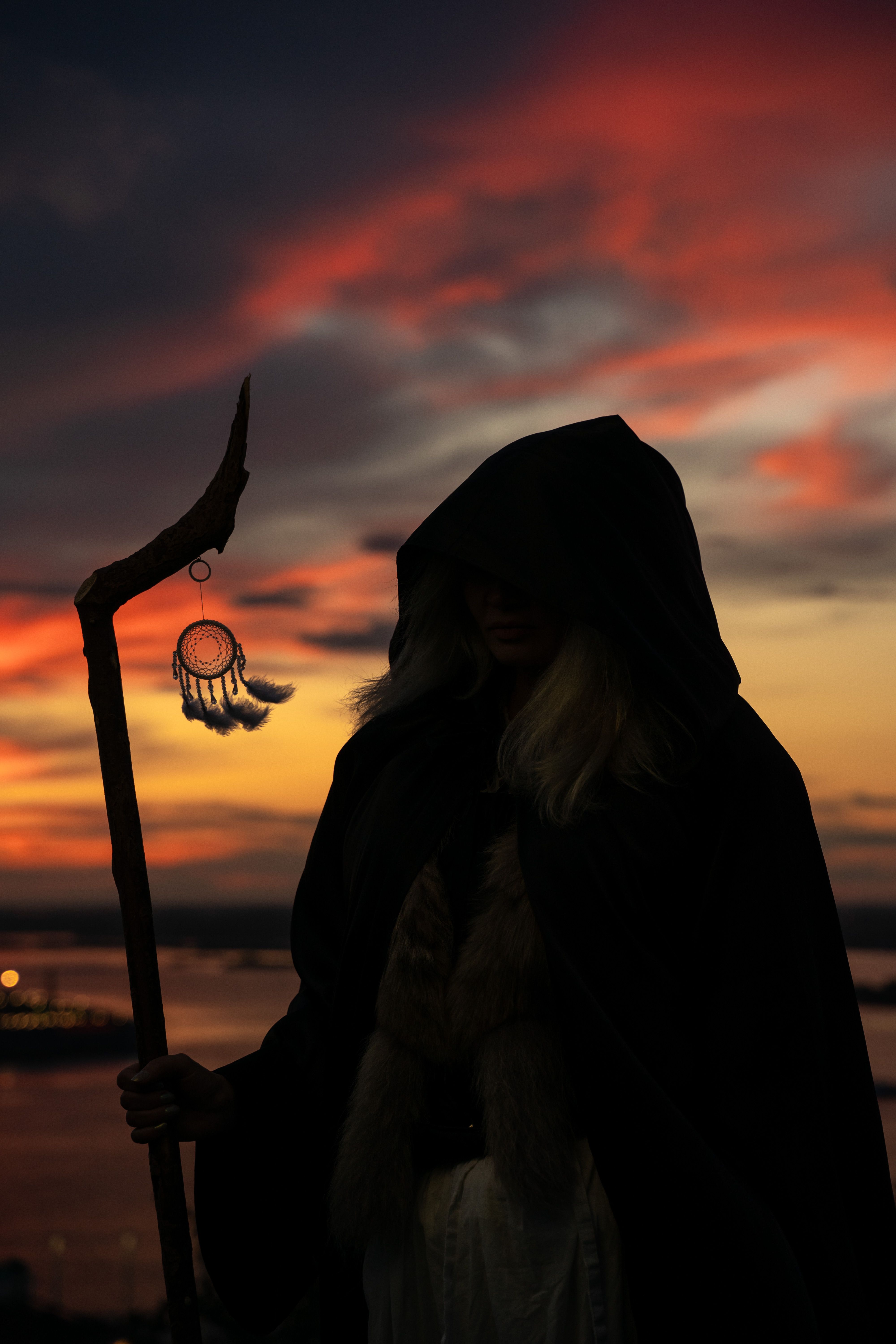 Mysterious hooded figure with staff silhouetted against vibrant sunset sky.