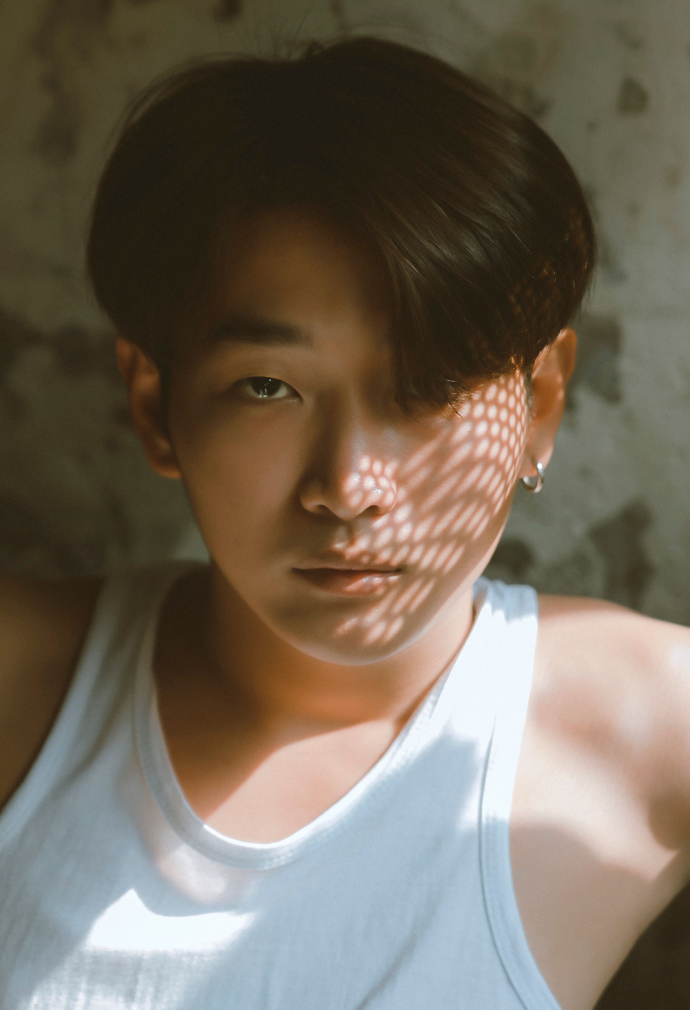 A pensive young man in a white tank top is illuminated by a textured shadow.