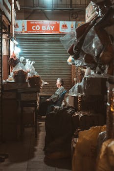 A vendor sits in a warmly lit indoor market stall, surrounded by packaged goods and a shuttered backdrop.