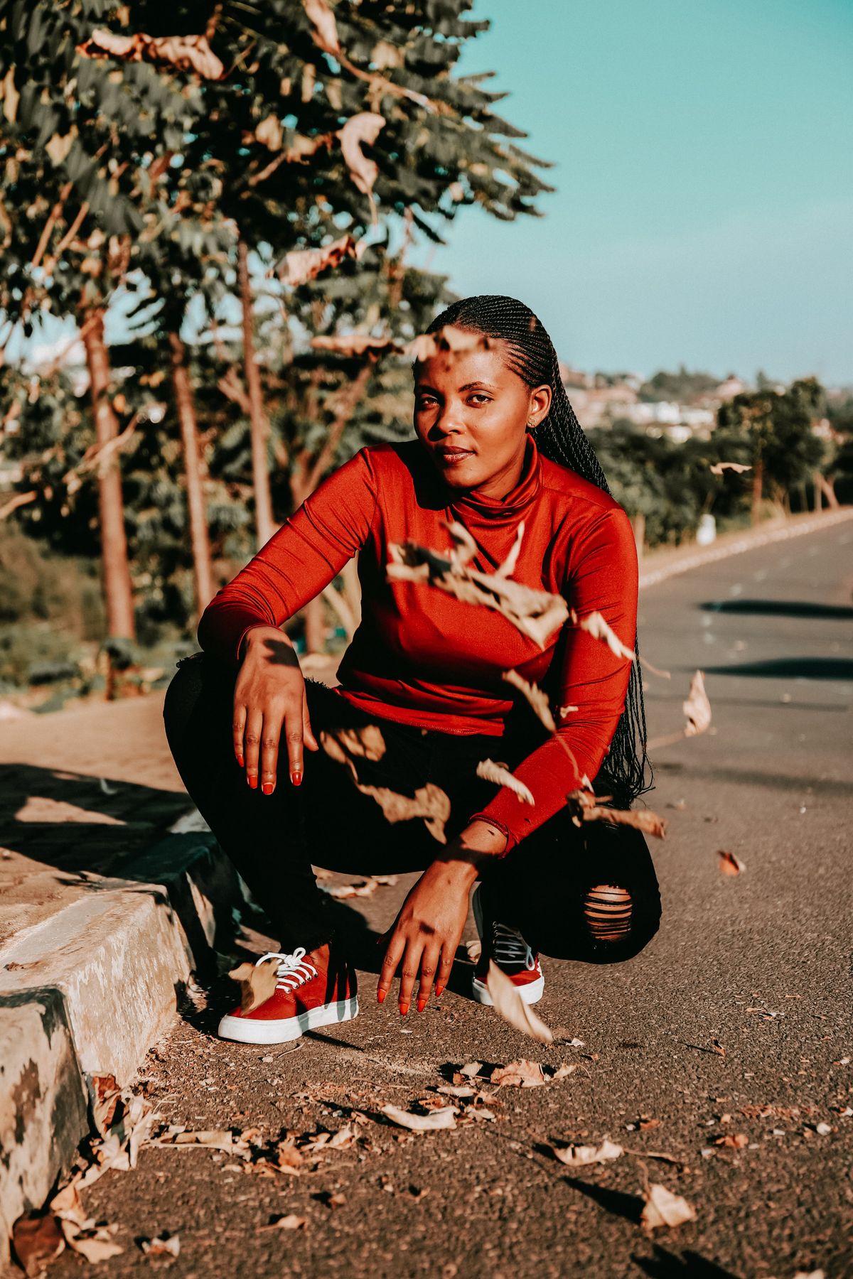 A confident woman in a red sweater poses outdoors amid falling leaves on a sunny Kigali street.