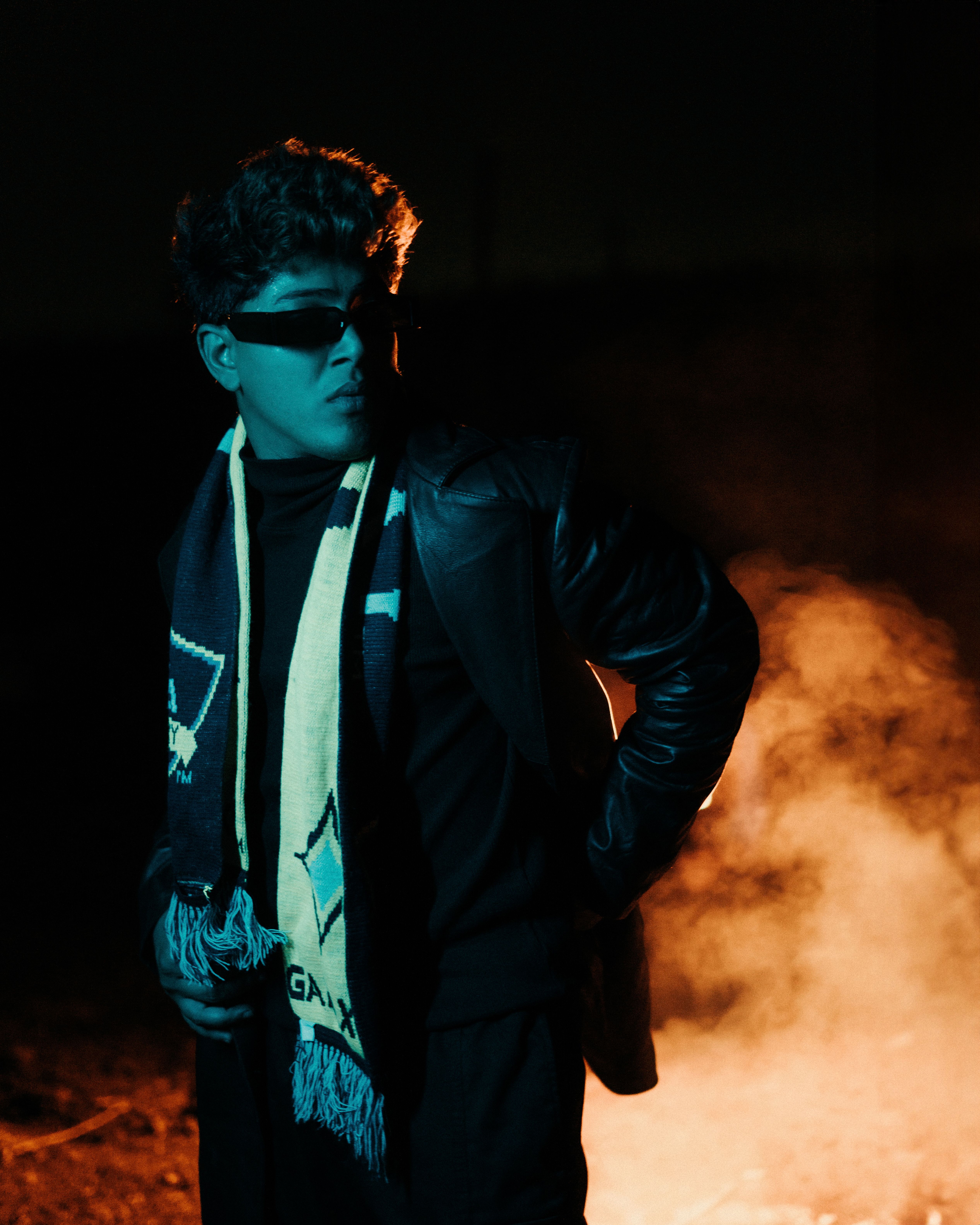 A fashionable man in sunglasses standing confidently against a dramatic smoky backdrop at night.