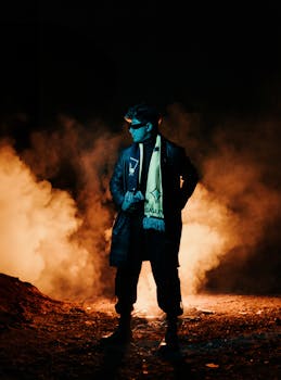 A man in sunglasses and a coat stands confidently with smoke glowing behind him at night.