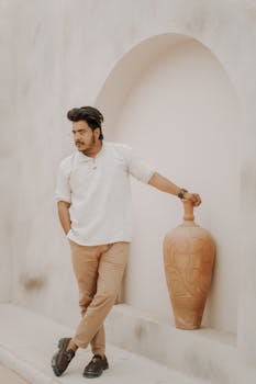 Fashionable man in smart casual wear poses with a clay vase in an elegant, minimalistic setting.