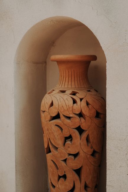 Artistic terracotta vase with detailed carvings displayed in an alcove, showcasing intricate craftsmanship.