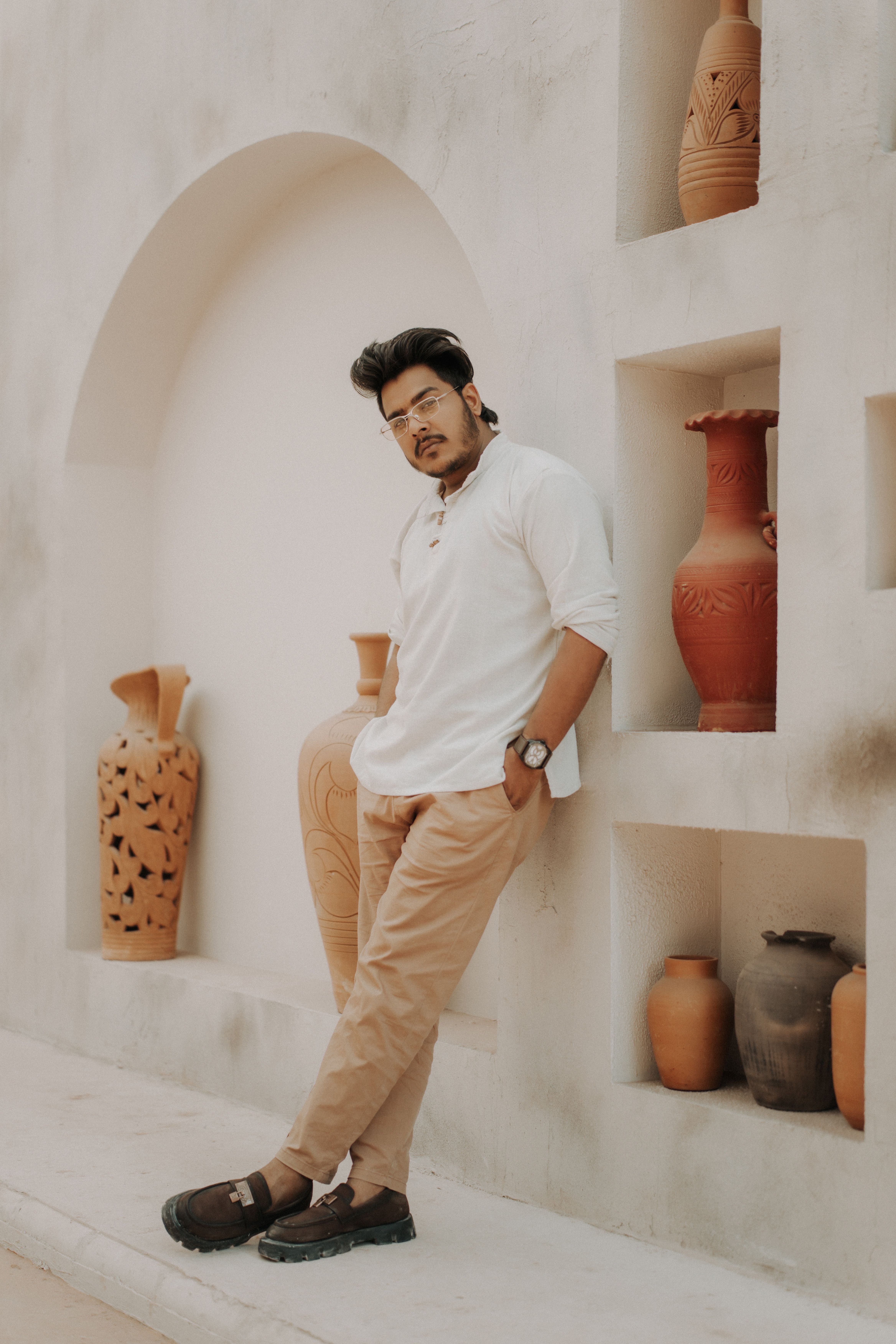 Man posing in white attire amidst rustic decor and clay vases indoors.