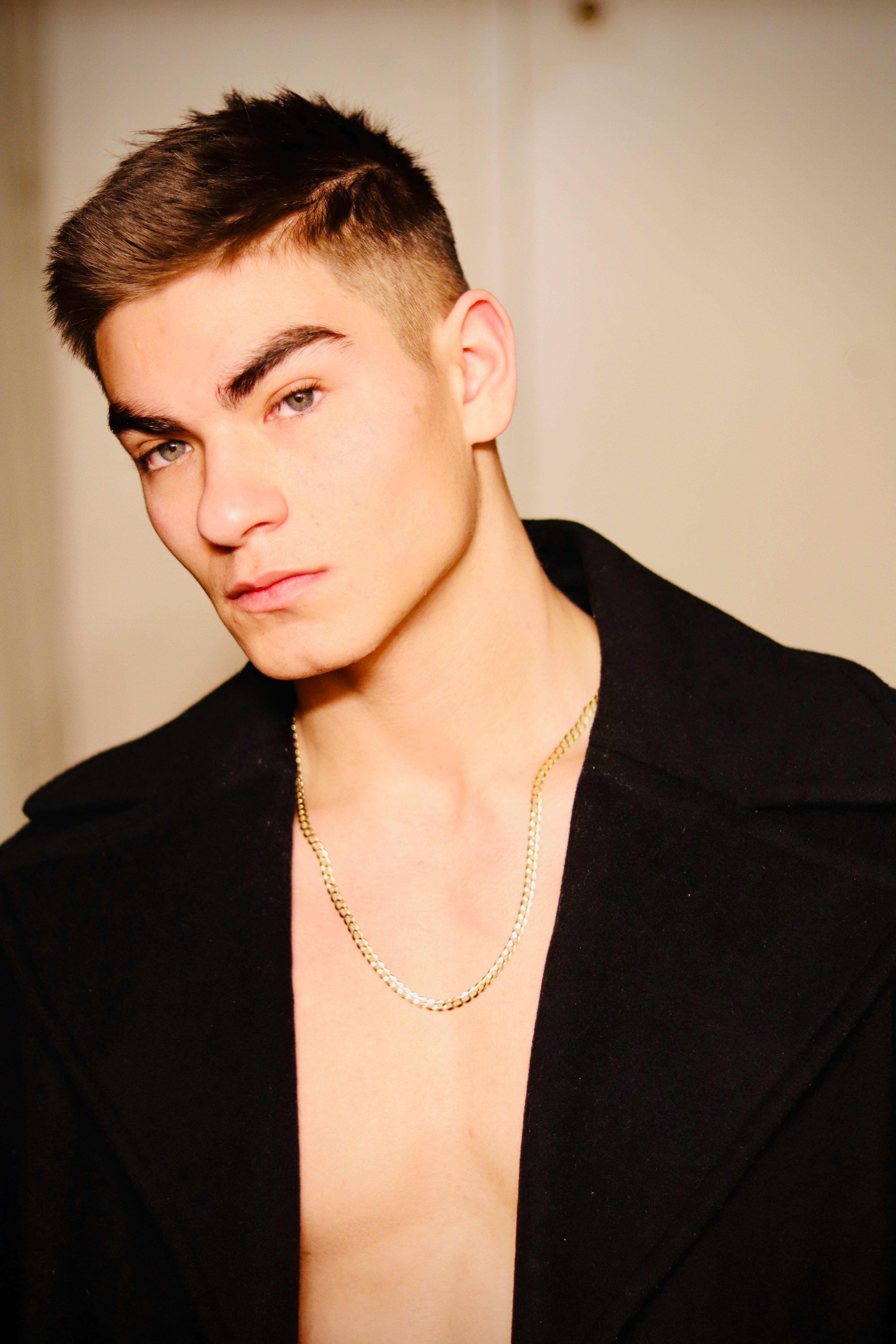 A stylish young male model wearing a black coat and gold chain, captured indoors.