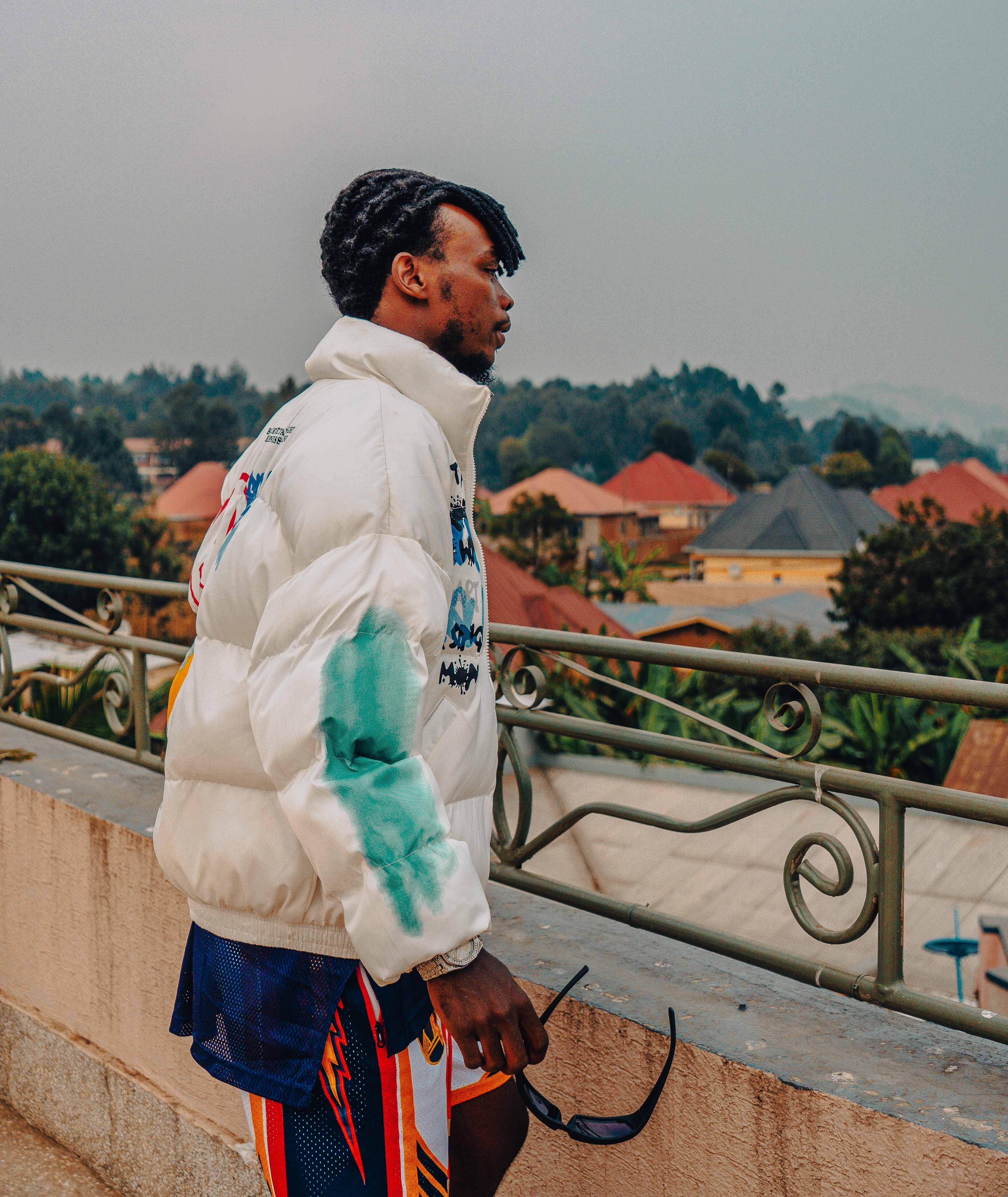 Stylishly dressed man in a white jacket gazing over cityscape from balcony.