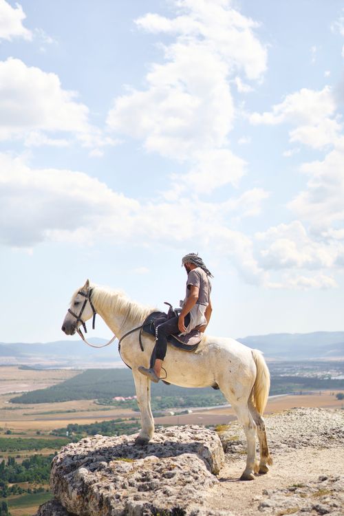Free A man rides a white horse atop a rocky cliff, surrounded by a vast scenic valley under a clear sky. Stock Photo
