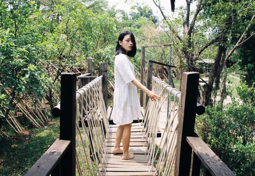 Free Woman in a white dress standing on a wooden bridge amidst lush greenery in a park Stock Photo