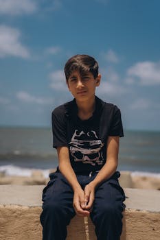 A young boy sits by the sea under a bright blue sky in Kafr El-Shaikh, Egypt.