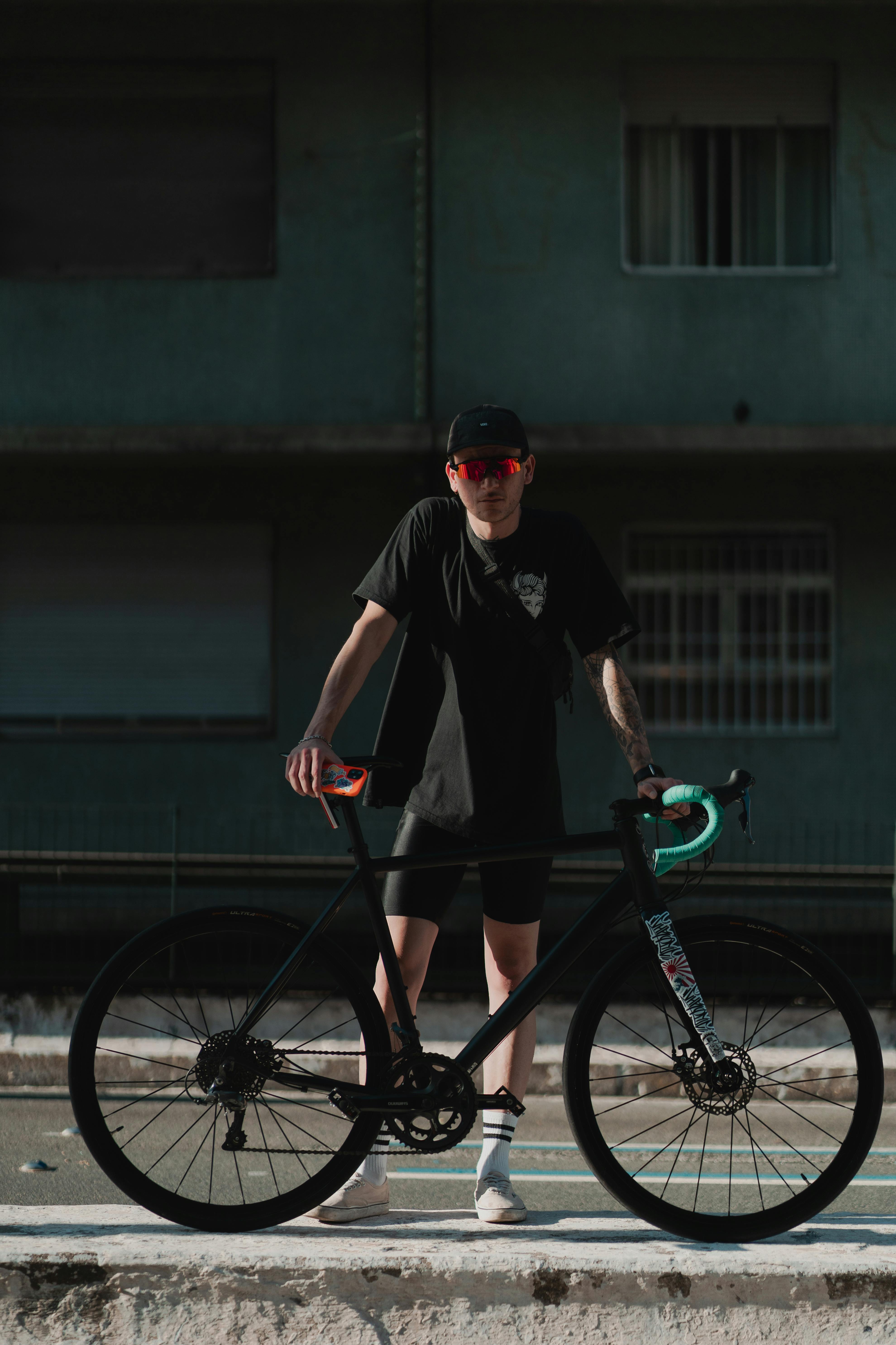 Man standing with a black bicycle in an urban setting on a sunny day.