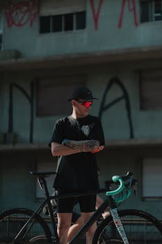 A trendy adult male cyclist poses with his bicycle against an urban backdrop.