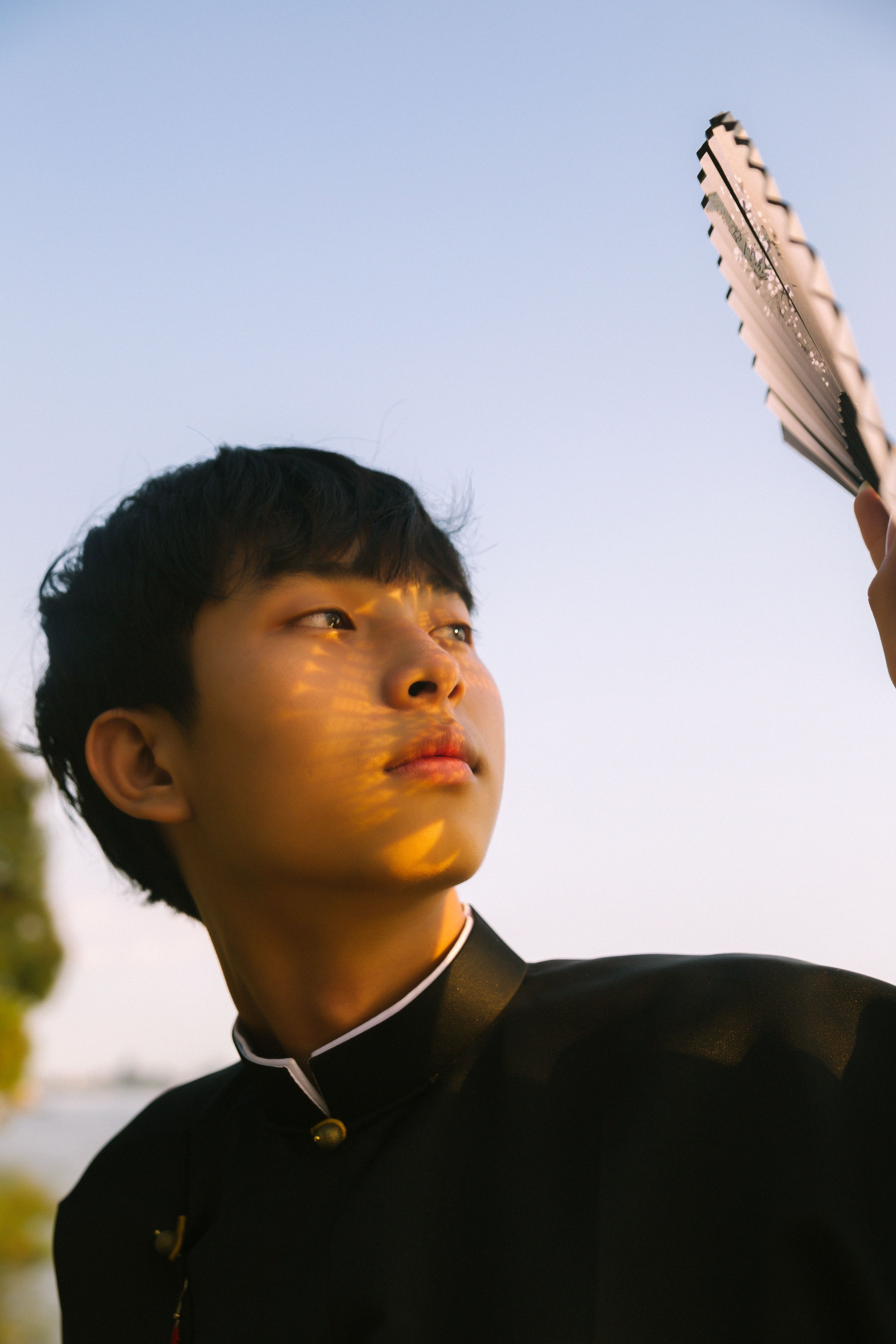 Portrait of a young man holding a hand fan outdoors during sunset with warm lighting.