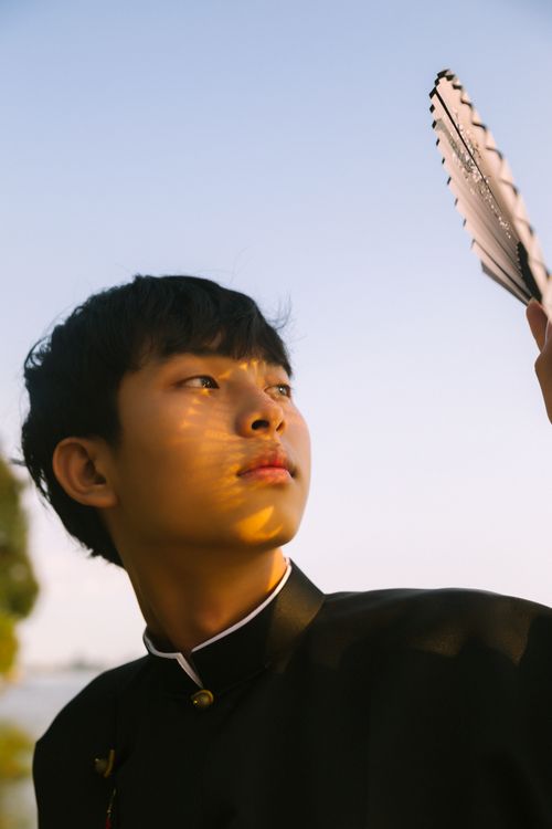 Free Portrait of a young man holding a hand fan outdoors during sunset with warm lighting. Stock Photo